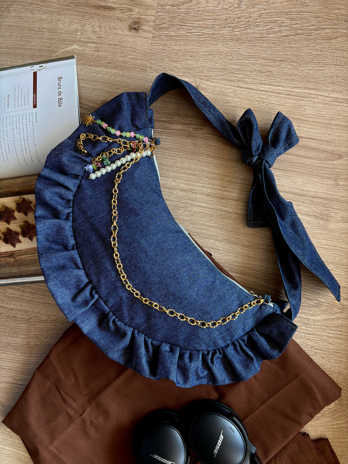 sac à main à froufrous Denim en jeans bleu foncé. posé sur une table en bois décorée d'une nappe en coton marron, un casque et un livre de recettes d'automne.