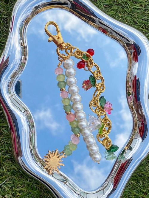 Bijou de sac fait main SIOU composé d'une chaîne dorée en acier inoxydable avec des breloques fleurs, cerises et tulipes accrochées, une chaines de perles nacrées ainsi qu'une chaines de perles en verre rose poudré, vert anis et vert sapin sur un petit miroir organique reflétant le ciel bleu du printemps avec de l'herbe autour.