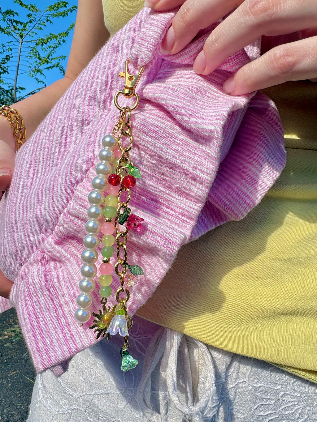 Bijou de sac fait main SIOU composé d'une chaîne dorée en acier inoxydable avec des breloques fleurs, cerises et tulipes accrochées, une chaines de perles nacrées ainsi qu'une chaines de perles en verre rose poudré, vert anis et vert sapin accroché au sac Tulipe de SIOU