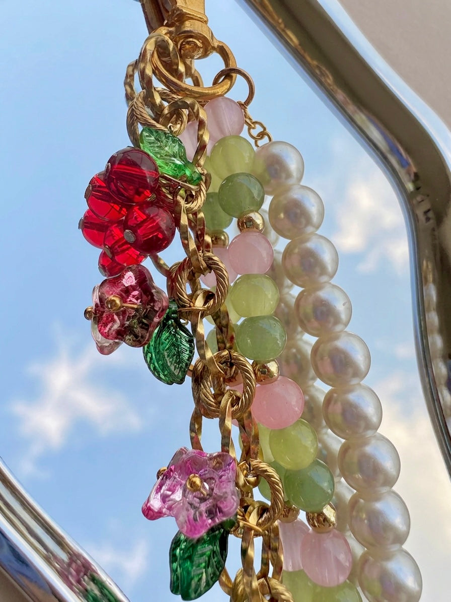 Plan rapproché du bijou de sac artisanal SIOU composé d'une chaîne dorée en acier inoxydable avec des breloques fleurs, cerises et tulipes accrochées, une chaines de perles nacrées ainsi qu'une chaines de perles en verre rose poudré, vert anis et vert sapin sur un petit miroir organique reflétant le ciel bleu du printemps avec de l'herbe autour.