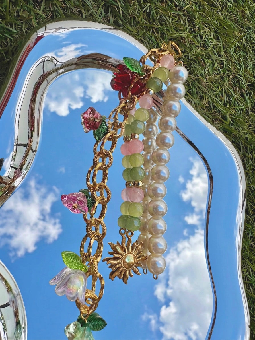 Bijou de sac assemblé à la main SIOU composé d'une chaîne dorée en acier inoxydable avec des breloques fleurs, cerises et tulipes accrochées, une chaines de perles nacrées ainsi qu'une chaines de perles en verre rose poudré, vert anis et vert sapin avec breloque vintage en acier inoxydable doré en forme de soleil sur un petit miroir organique reflétant le ciel bleu du printemps avec de l'herbe autour.