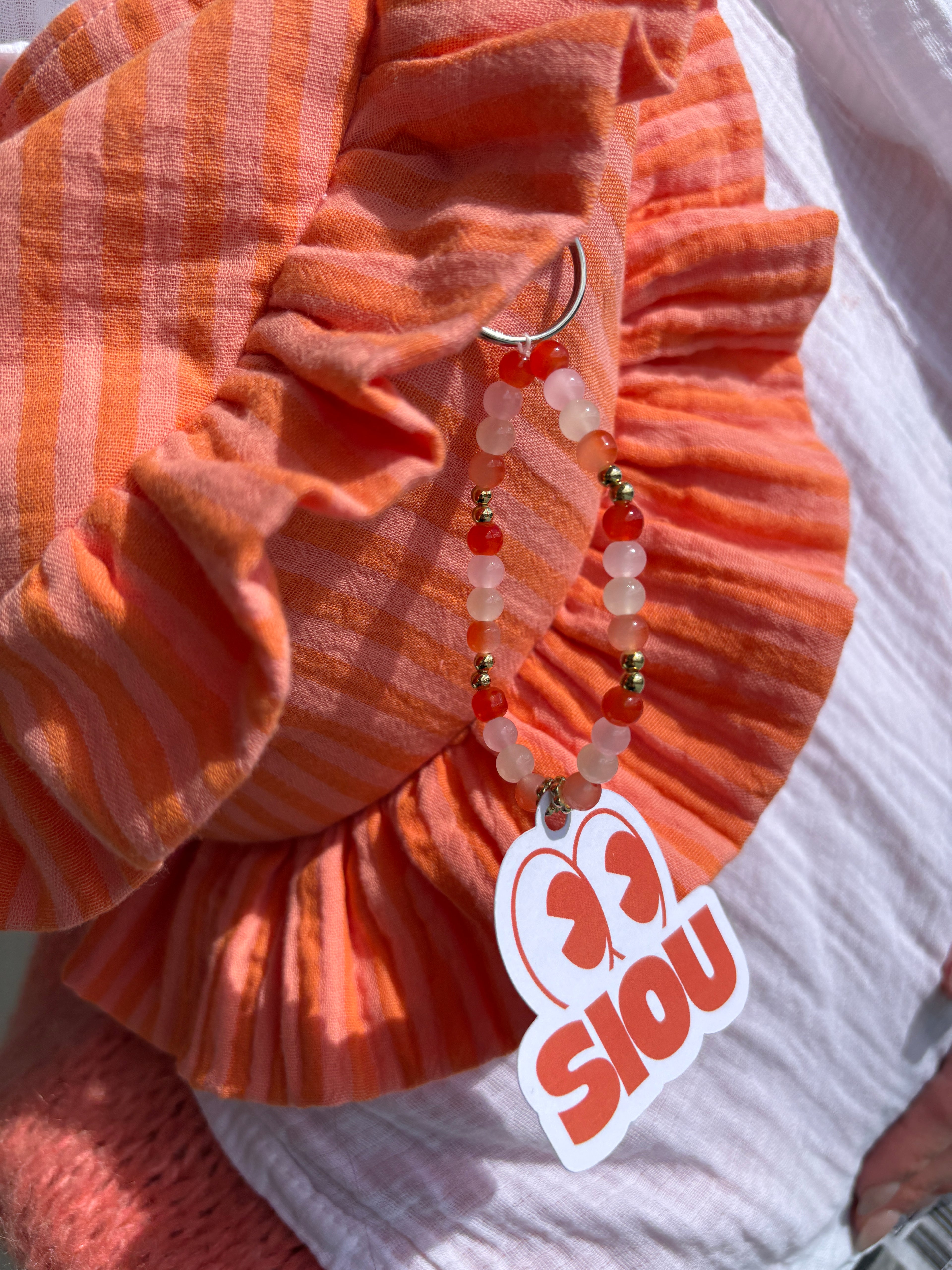 Bijou de sac fait main en perles de verre couleur orange, rose poudré, beige, doré et argenté avec l'étiquette de la marque SIOU du sac à main en tissu gaze de coton à volants en forme de croissant, cousu main, coloris rayures orange et rose Pêche-Abricot, collection Printemps, sur une nappe de pique-nique, porté par une femme habillé d'une chemise blanche en gaze de coton.