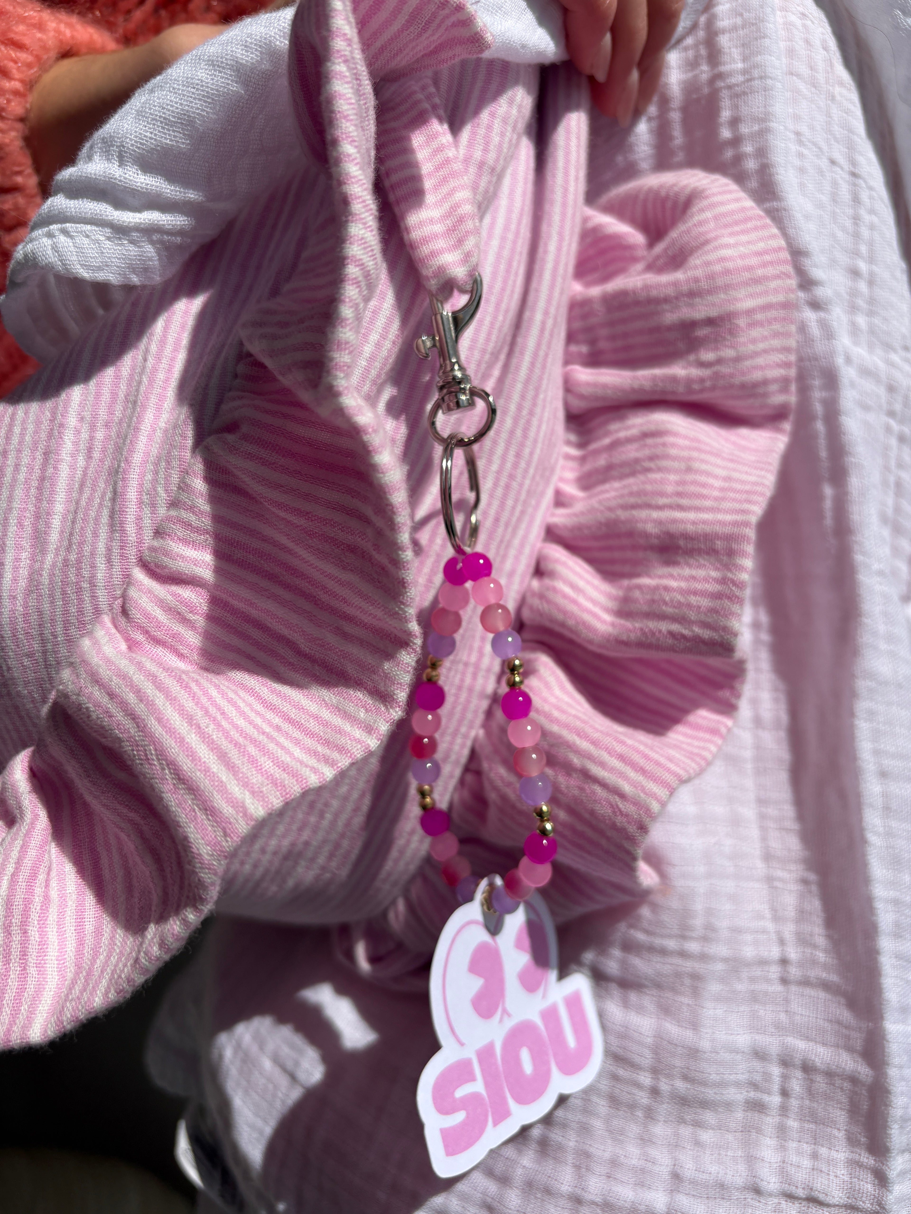 Bijou de sac en perle de verre fait main couleur rose fuchsia, rose poudré, violet lilas et doré et argenté avec l'étiquette de la marque SIOU du sac à main en tissu gaze de coton à volants en forme de croissant, coloris rayures rose et blanc Tulipe, collection Printemps, porté à l'épaule par une femme portant une chemise blanche en gaze de coton