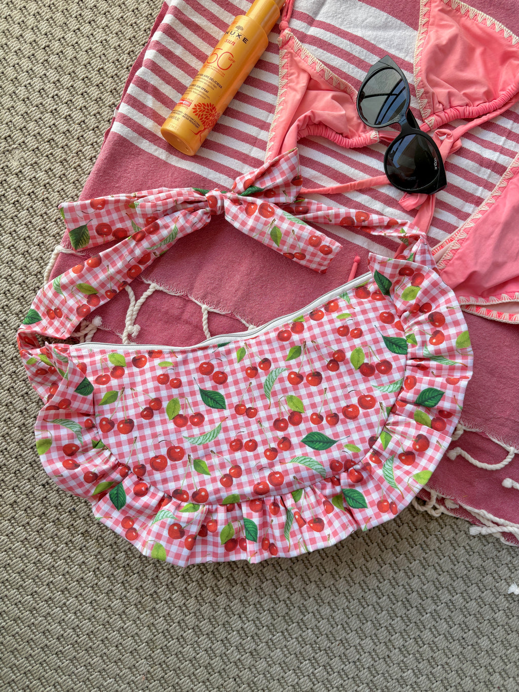 Sac à main en tissu à volants en forme de demi-lune/croissant, cousu main, coloris en vichy rose et blanc avec des illustrations de cerises, collection
Été, sur une serviette de plage entouré d'un maillot de bain deux pièces rose, une crème solaire et des lunettes de soleil.