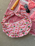 Sac à main en tissu à volants en forme de demi-lune/croissant, cousu main, coloris en vichy rose et blanc avec des illustrations de cerises, collection
Été, sur une serviette de plage entouré d'un maillot de bain deux pièces rose, une crème solaire et des lunettes de soleil.