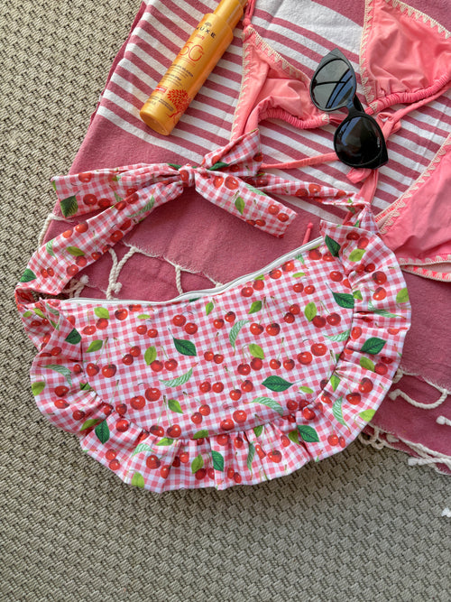 Sac à main en tissu à volants en forme de demi-lune/croissant, cousu main, coloris en vichy rose et blanc avec des illustrations de cerises, collection
Été, sur une serviette de plage entouré d'un maillot de bain deux pièces rose, une crème solaire et des lunettes de soleil.