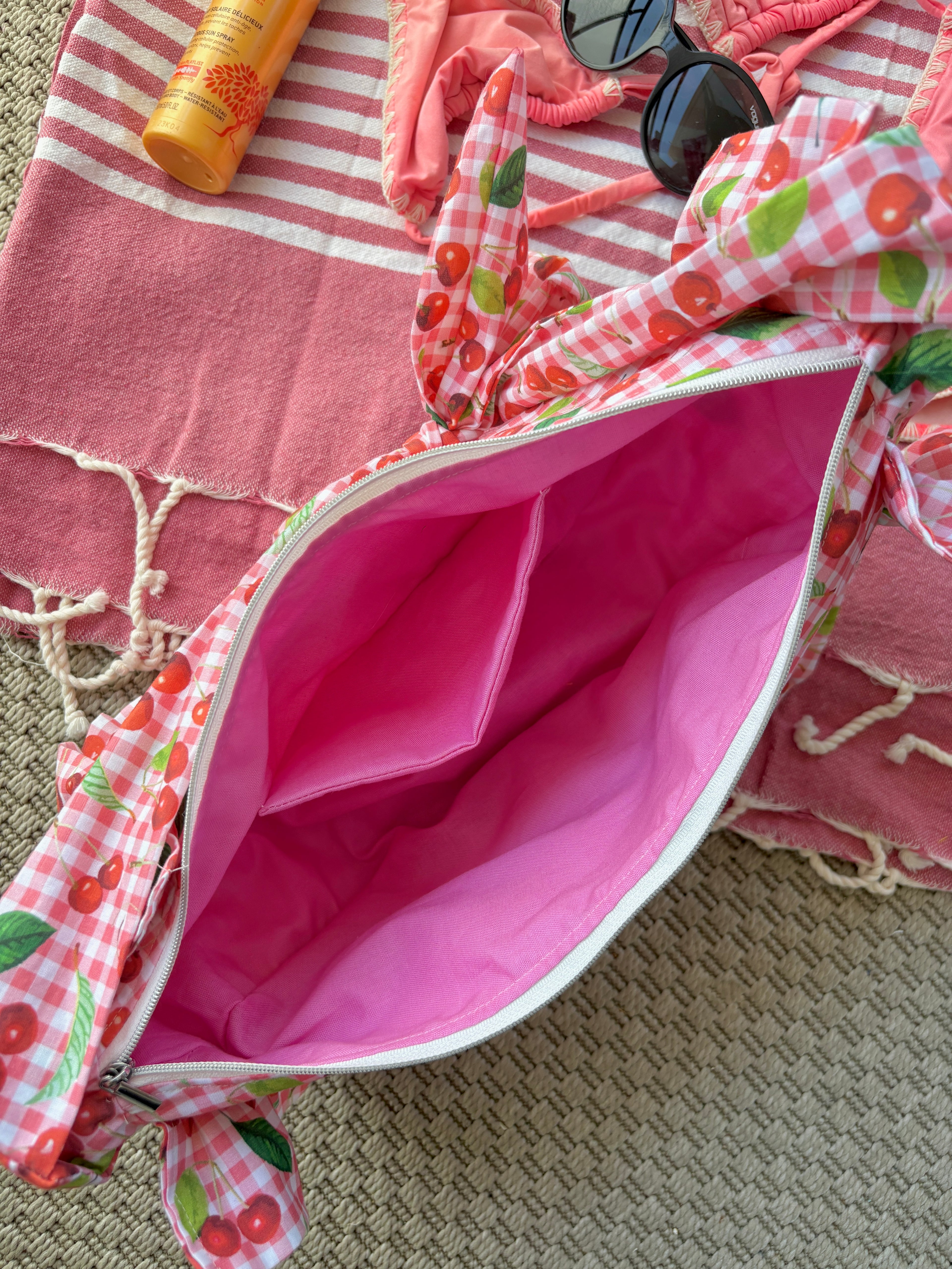 Intérieur rose du sac à main vu du dessus en tissu à volants en forme de demi-lune/croissant, cousu main, coloris en vichy rose et blanc avec des illustrations de cerises, collection
Été, sur une serviette de plage entouré d'un maillot de bain deux pièces rose, une crème solaire et des lunettes de soleil.