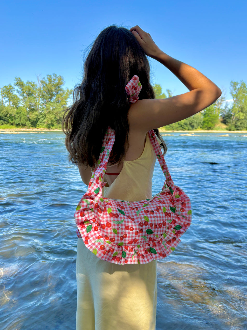 Jeune femme habillée d'une longue robe jaune pastel portant à l'épaule le sac à main Cerise de SIOU cousu main en forme de croissant à volants. Son tissu est composé de cerises sur un fond vichy vichy rose et blanc.