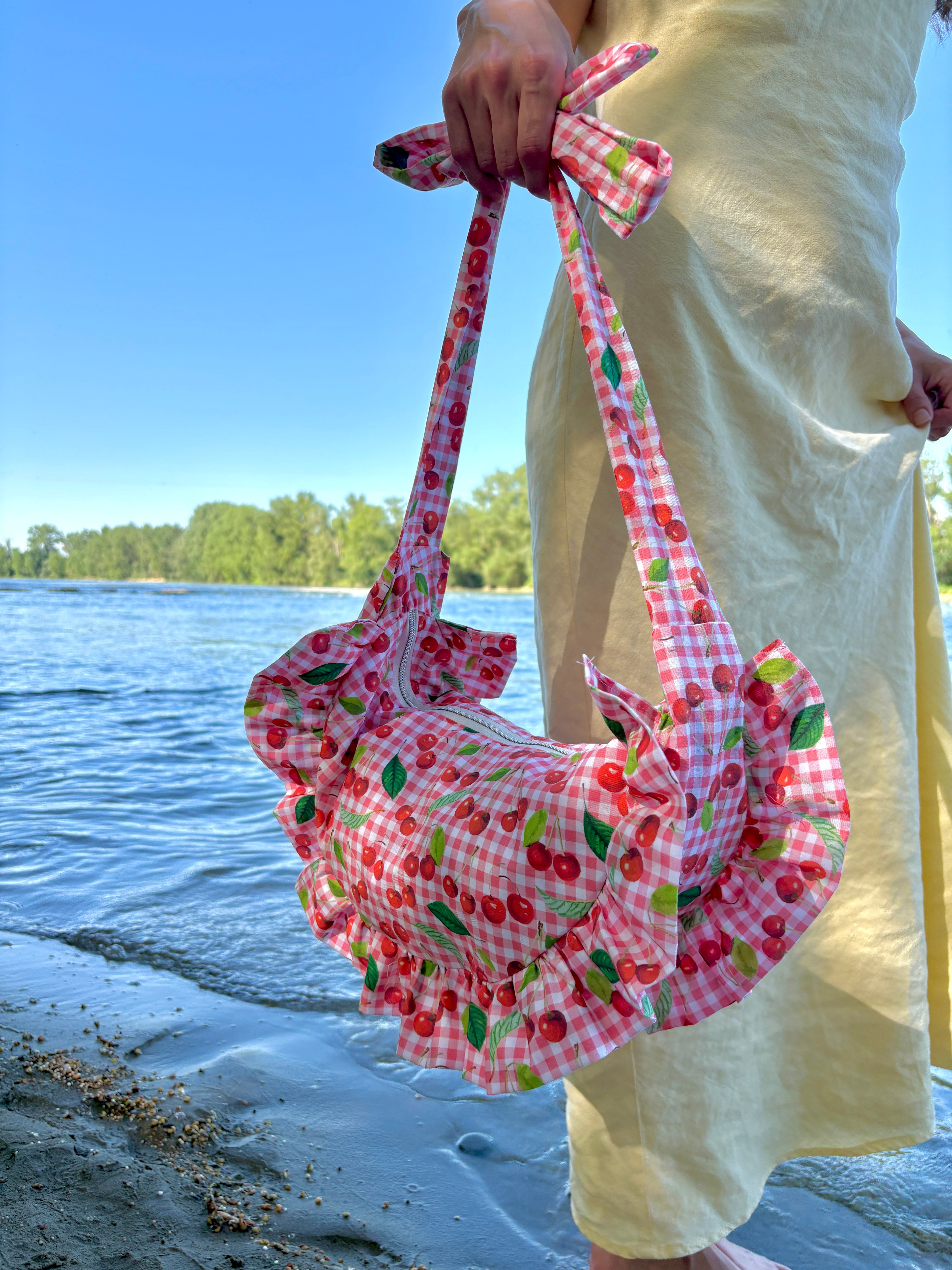 Jeune femme, marchant au bord de l'eau habillée d'une robe longue jaune poussin portant à la main, le long de son corps, le sac Cerise de SIOU dont le motif du tissu est composé de cerises sur un fond vichy rose et blanc.
