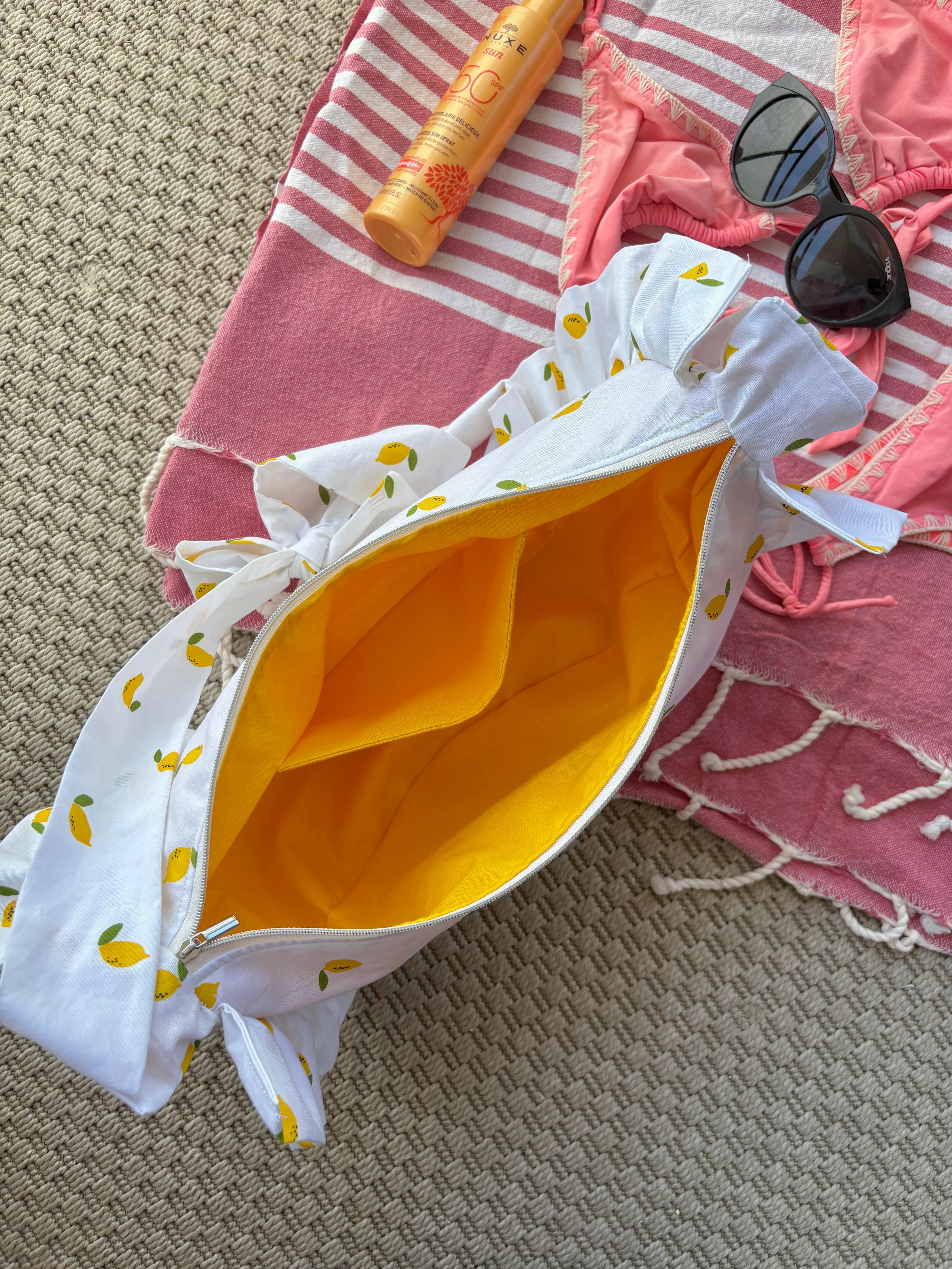 Sac à main en tissu à volants en forme de demi-lune/croissant, cousu main, coloris mini citrons sur fond blanc la doublure intérieure est jaune soleil, collection
Été, sur une serviette de plage, sur du sable avec un bikini, un tube de crème solaire et des lunettes de soleil autour.