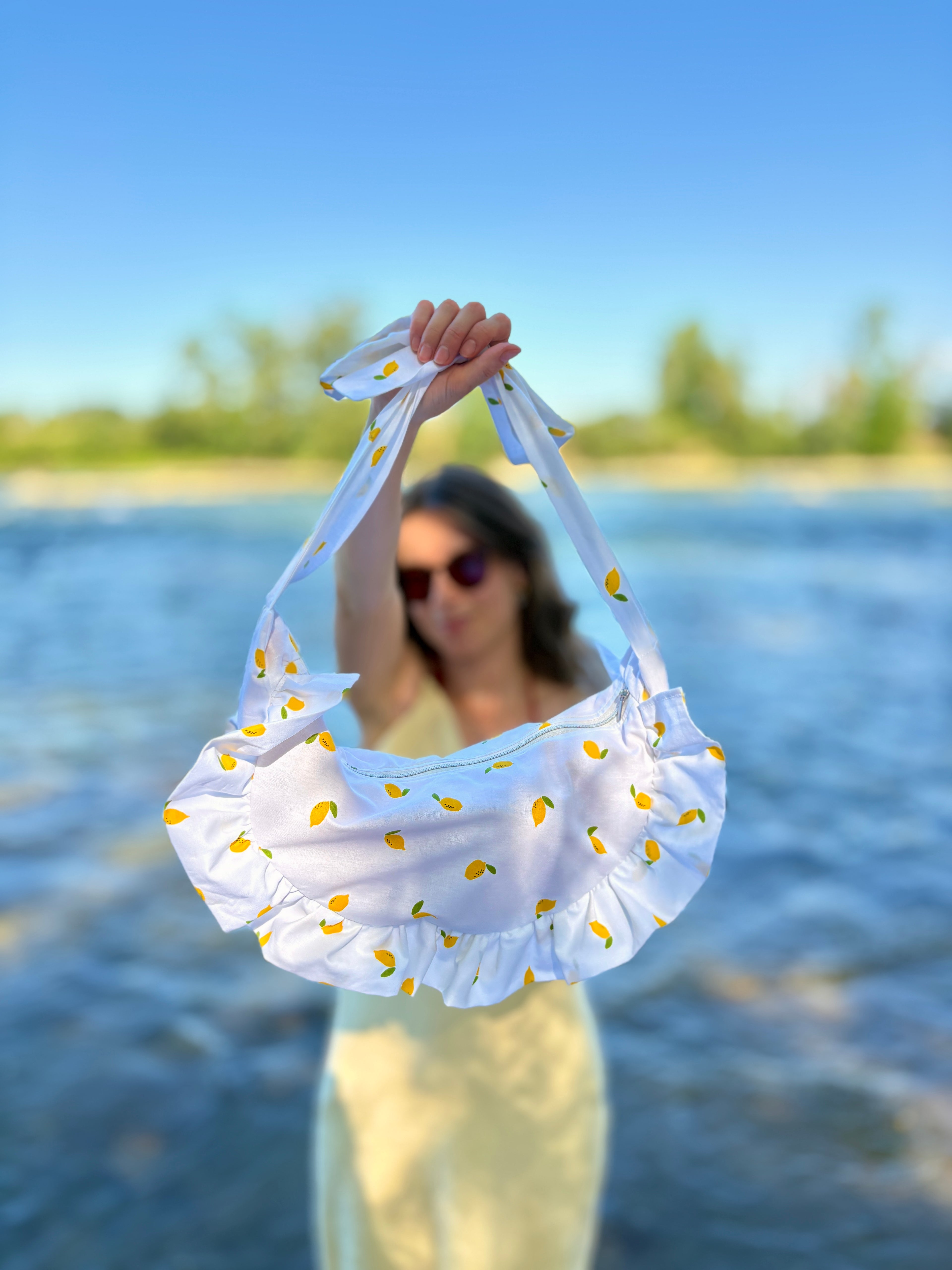 jeune femme portant à bout de bras le sac SIOU Citron, un sac en tissu au motif de mini citrons sur fond blanc. en arrière plan, une étendue d'eau illuminée par les reflets du soleil.
