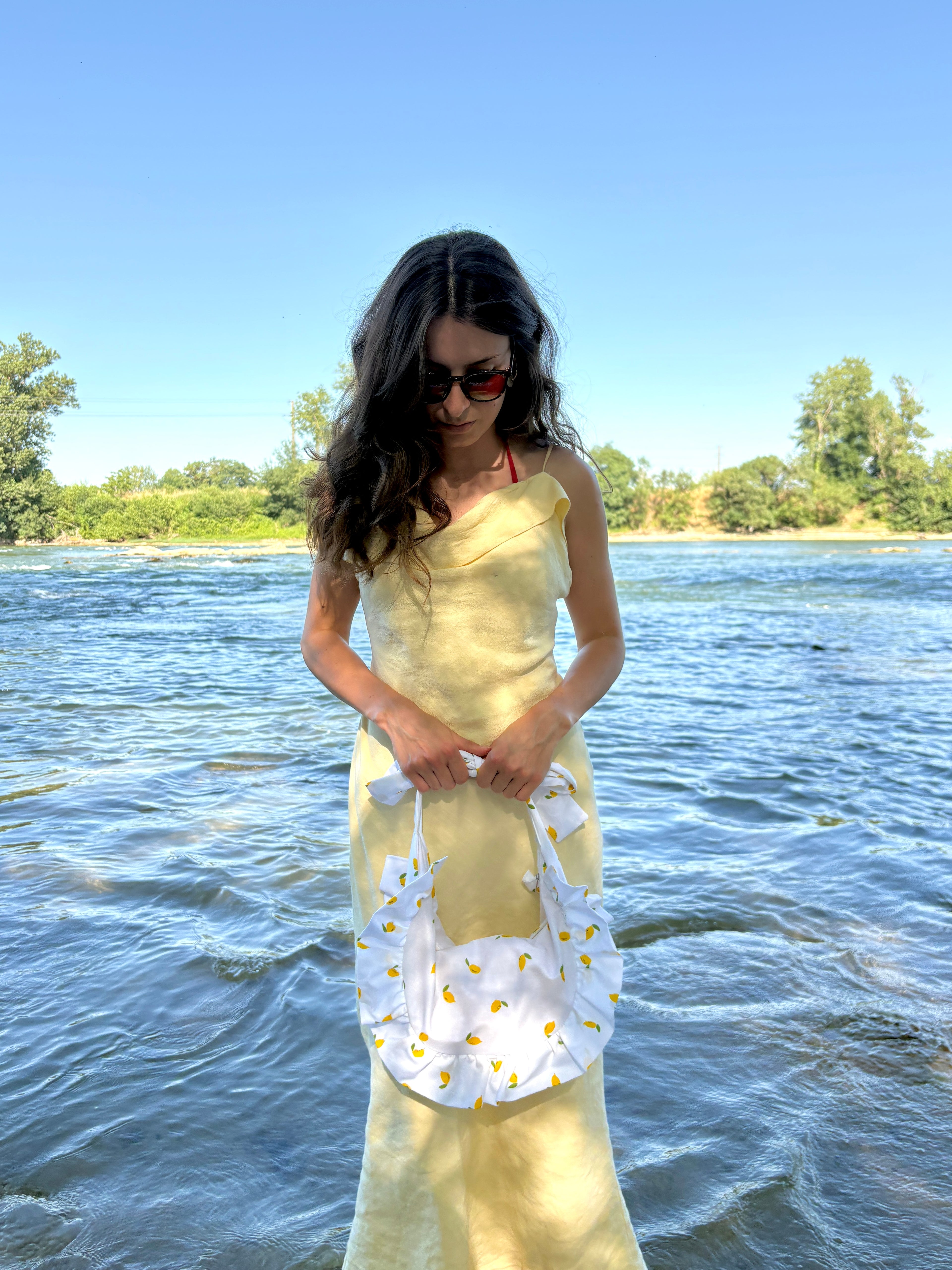 jeune femme portant à bout de bras, au niveau de ses genoux, le sac SIOU Citron, un sac en tissu au motif de mini citrons sur fond blanc. en arrière plan, une étendue d'eau illuminée par les reflets du soleil.