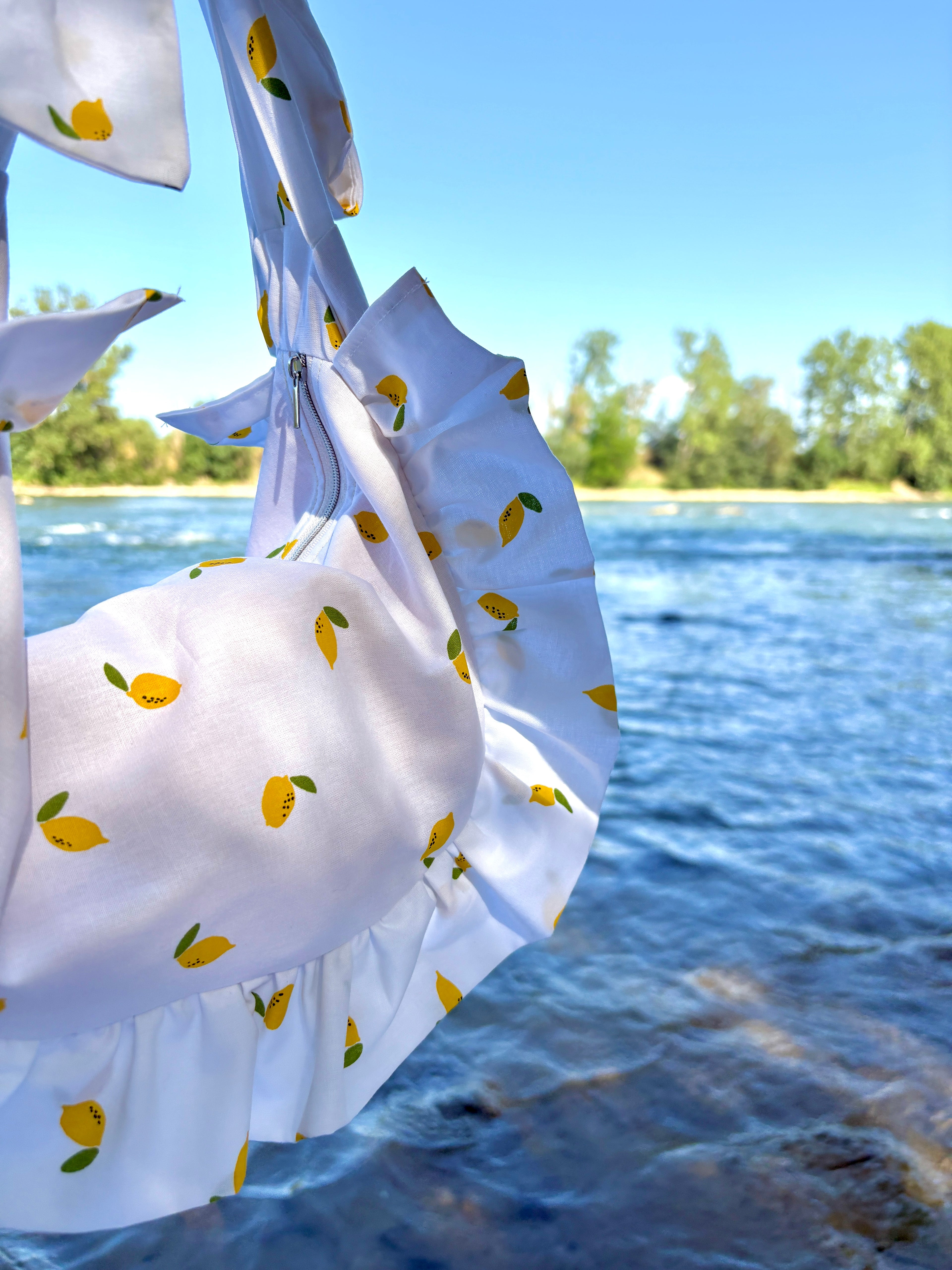 Zoom sur le tissu du sac à main SIOU Citron, des mini citrons sur fond blanc. un sac demi-lune à froufrous avec en arrière plan une étendue d'eau illuminée par les reflets du soleil.