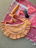Sac à main Corail de SIOU en tissu à volants en forme de demi-lune/croissant, cousu main, coloris en vichy rose et jaune, collection
Été, sur une serviette de plage entouré d'un maillot de bain deux pièces rose, une crème solaire et des lunettes de soleil.