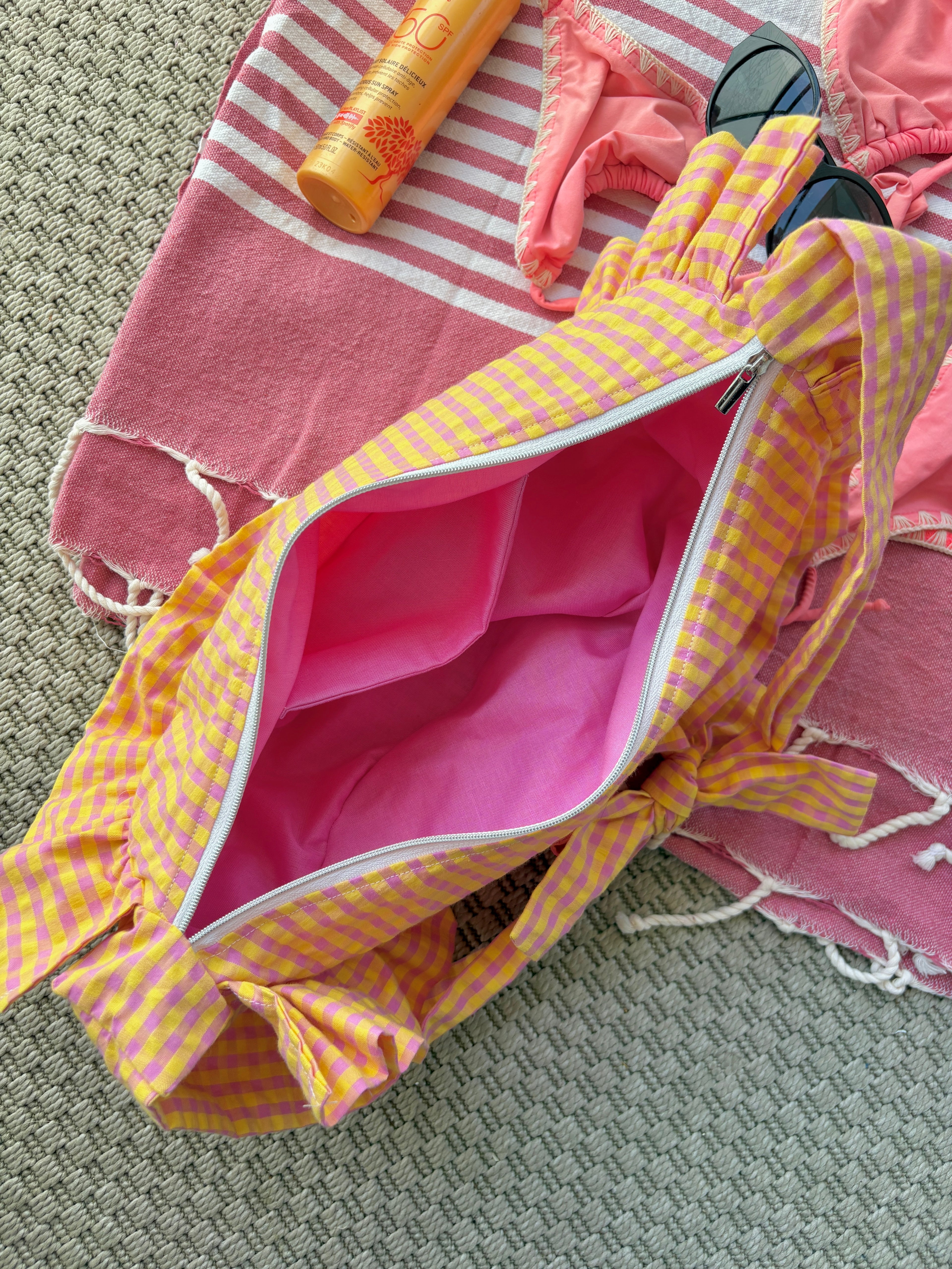 Vue du dessus de l'intérieur rose du sac fait à la main Corail de la marque SIOU dont le tissus a un motif vichy rose et jaune. Le sac en forme de demi-lune à froufrous/volants est posé sur une serviette de plage rose avec un bikini rose, un tube de crème solaire et des lunettes de soleil autour.