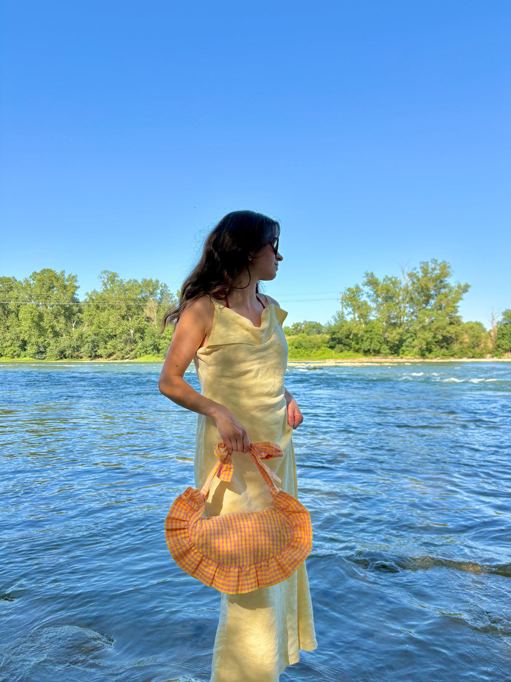 Jeune femme au bord de l'eau sur le sable vêtue d'une jolie robe longue jaune porte, à la main, le sac Corail de la marque SIOU au tissu vichy rose et jaune. 