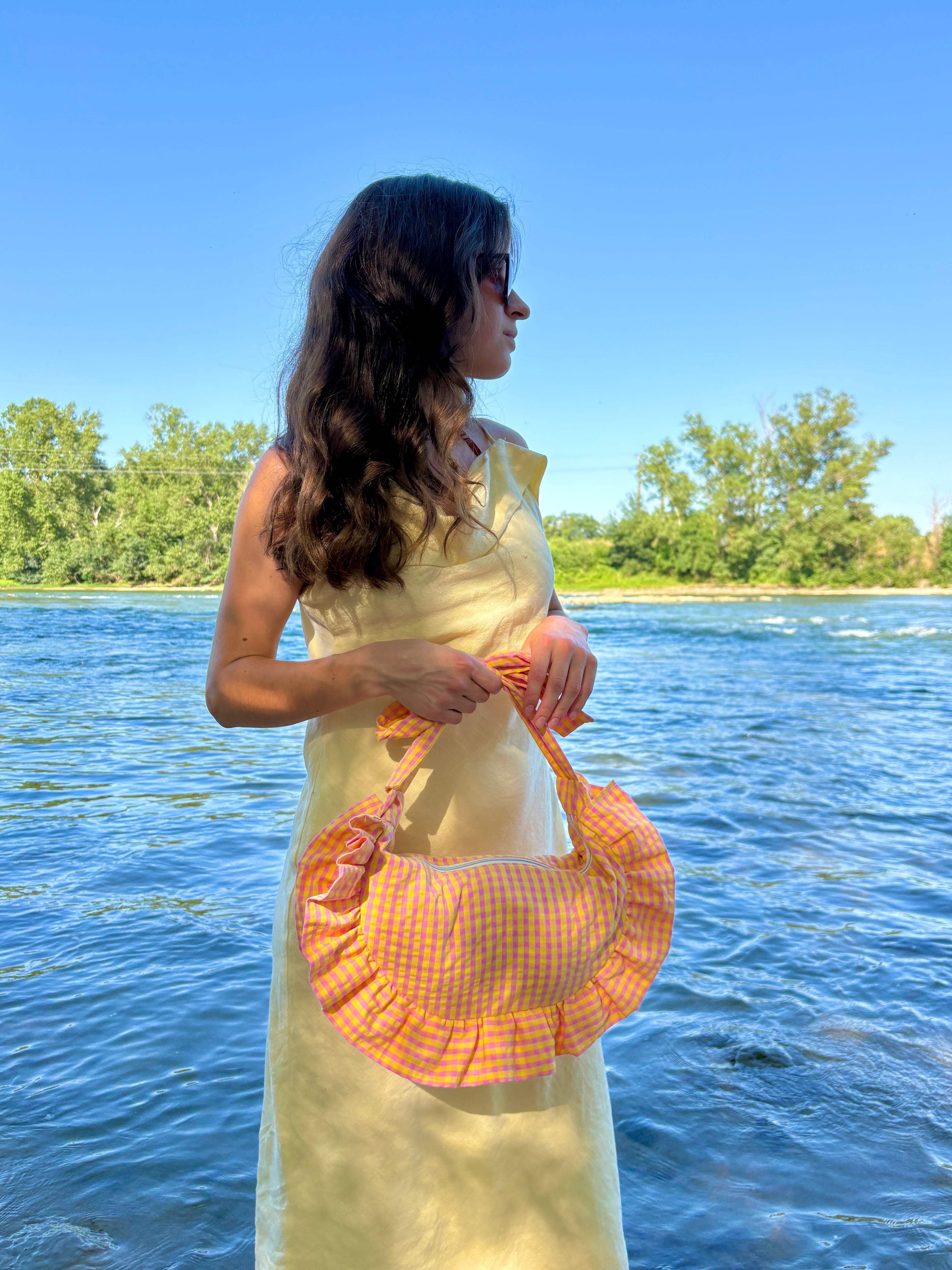 Femme portant à ses main le sac cousu main Corail de la marque SIOU, ses anses sont nouées, le tissu est un vichy rose et jaune. Le sac demi-lune/croissant porte des froufrous autour. Le décor est une plage avec de l'eau bleue et des arbres en arrière-plan.