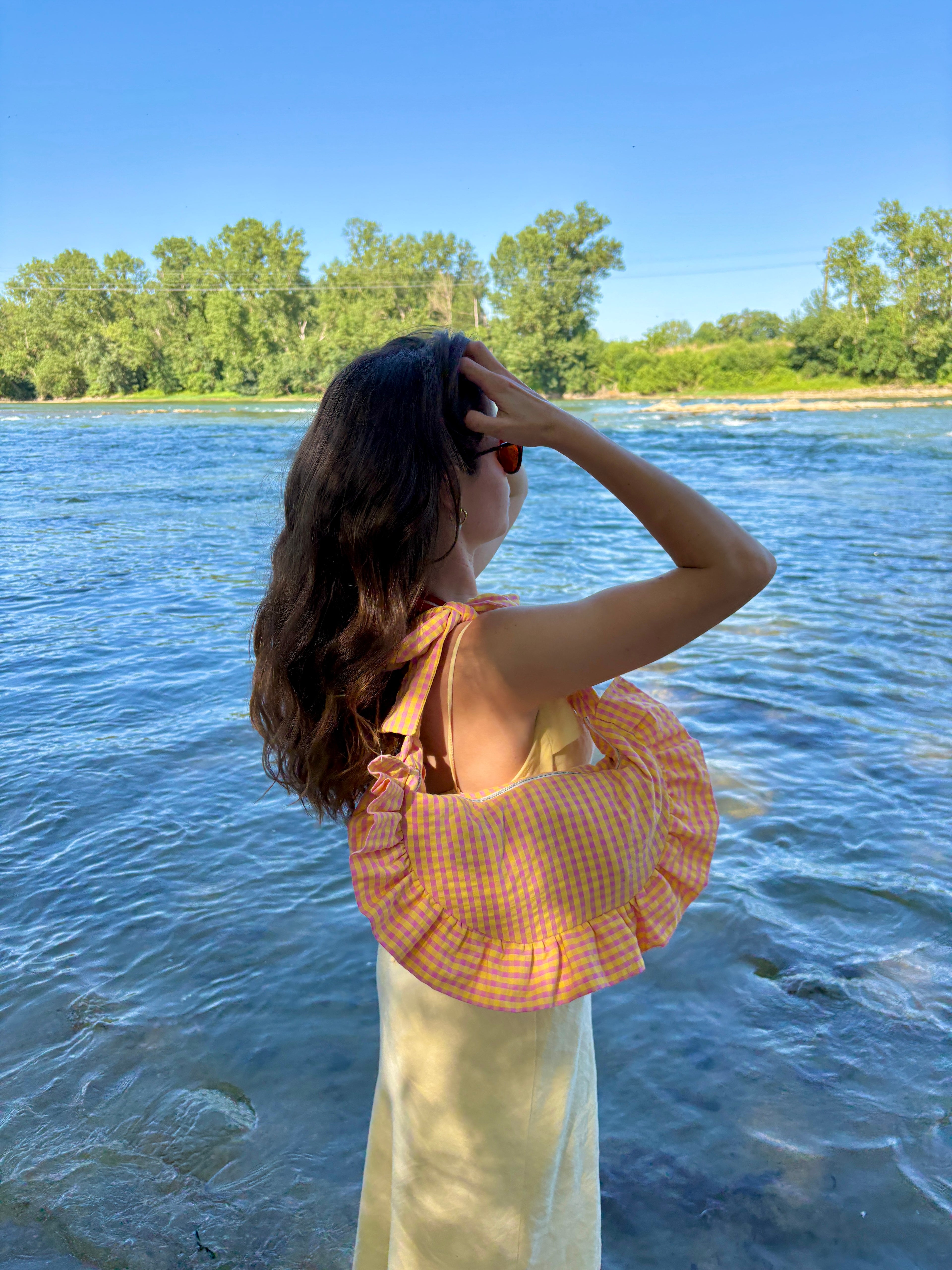 femme au bord de l'eau vêtue d'une longue robe jaune pastel et du sac cousu main Corail de la marque SIOU qui a une forme de demi-lune/croissant avec des volants/froufrous, passe les mains dans ses cheveux.