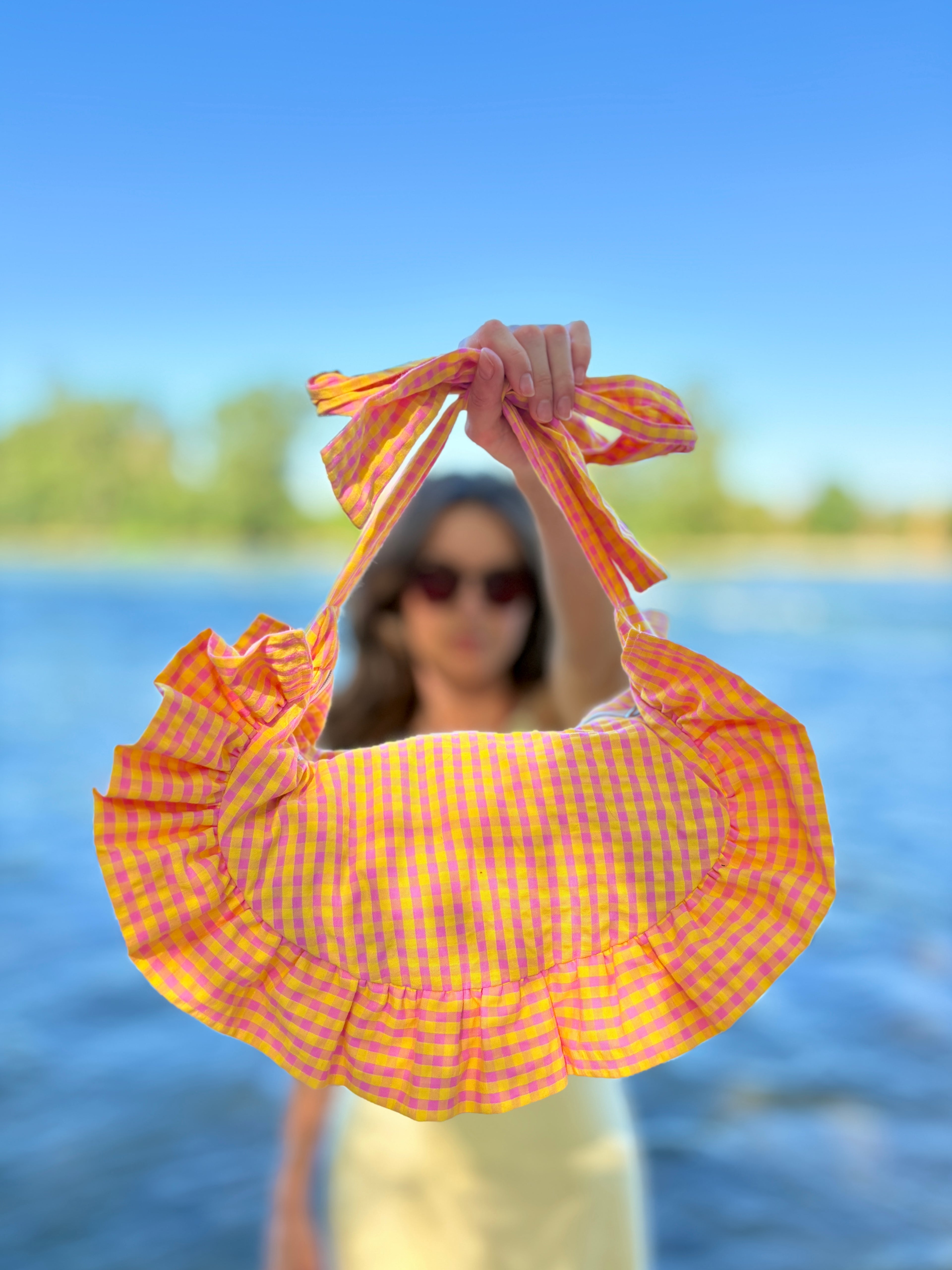 Effet portrait du sac cousu main Corail, en vichy rose et jaune de la marque SIOU, c'est un sac à main où l'on peut nouer les anses pour capter la taille de ses anses et le porter soit en bandoulière soit à la main. Le sac, en forme de croissant arborant des volants, est présenté au premier plan par une jeune femme qui est à l'arrière plan au bord de l'eau.