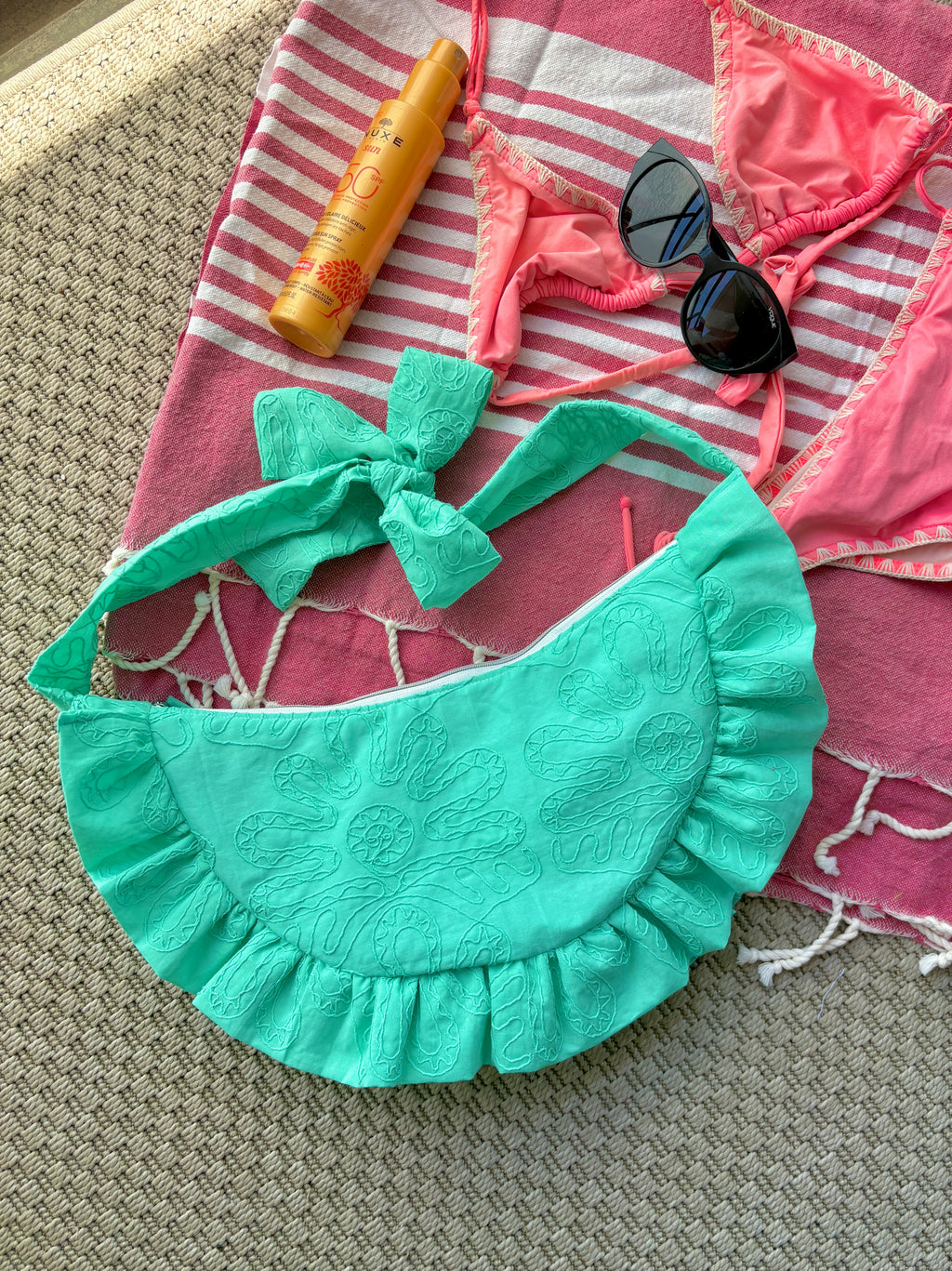 Sac à main Lagon de la marque SIOU en tissu à volants en forme de demi-lune/croissant, cousu main, coloris turquoise avec des broderies en forme de fleur, collection Été, sur une serviette de plage entouré d'un maillot de bain deux pièces rose, une crème solaire et des lunettes de soleil.