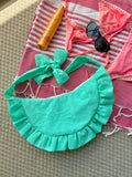 Sac à main Lagon de la marque SIOU en tissu à volants en forme de demi-lune/croissant, cousu main, coloris turquoise avec des broderies en forme de fleur, collection Été, sur une serviette de plage entouré d'un maillot de bain deux pièces rose, une crème solaire et des lunettes de soleil.