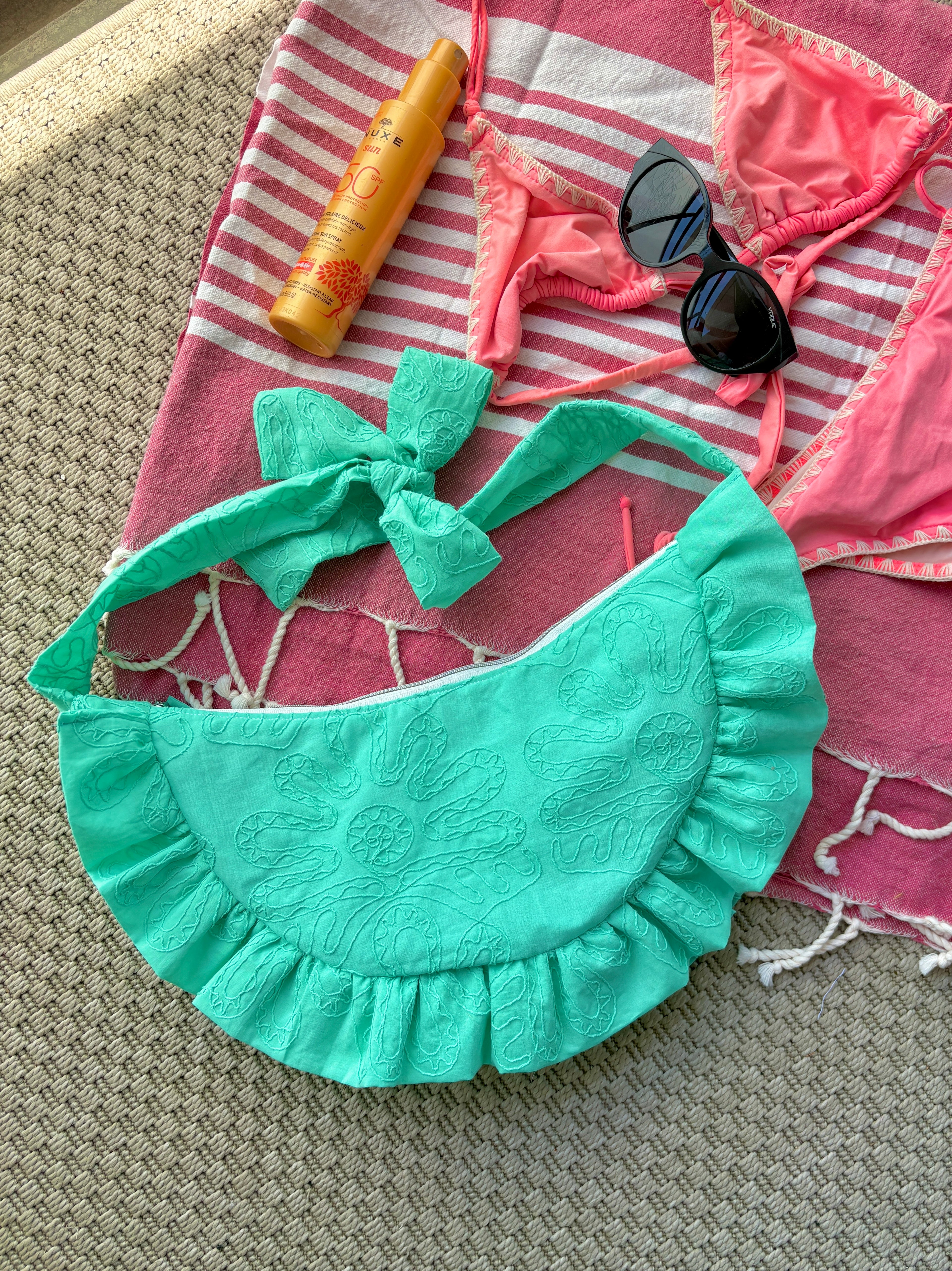 Sac à main Lagon de la marque SIOU en tissu à volants en forme de demi-lune/croissant, cousu main, coloris turquoise avec des broderies en forme de fleur, collection Été, sur une serviette de plage entouré d'un maillot de bain deux pièces rose, une crème solaire et des lunettes de soleil.