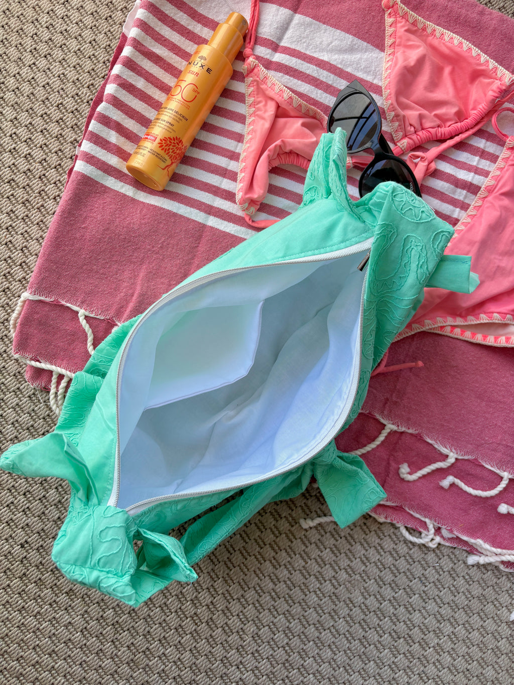 Vue du dessus de l'intérieur blanc du sac à main Lagon de la marque SIOU en tissu à volants en forme de demi-lune/croissant, cousu main, coloris turquoise avec des broderies en forme de fleur, collection Été, sur une serviette de plage rose, entouré d'un maillot de bain deux pièces rose, une crème solaire et des lunettes de soleil.