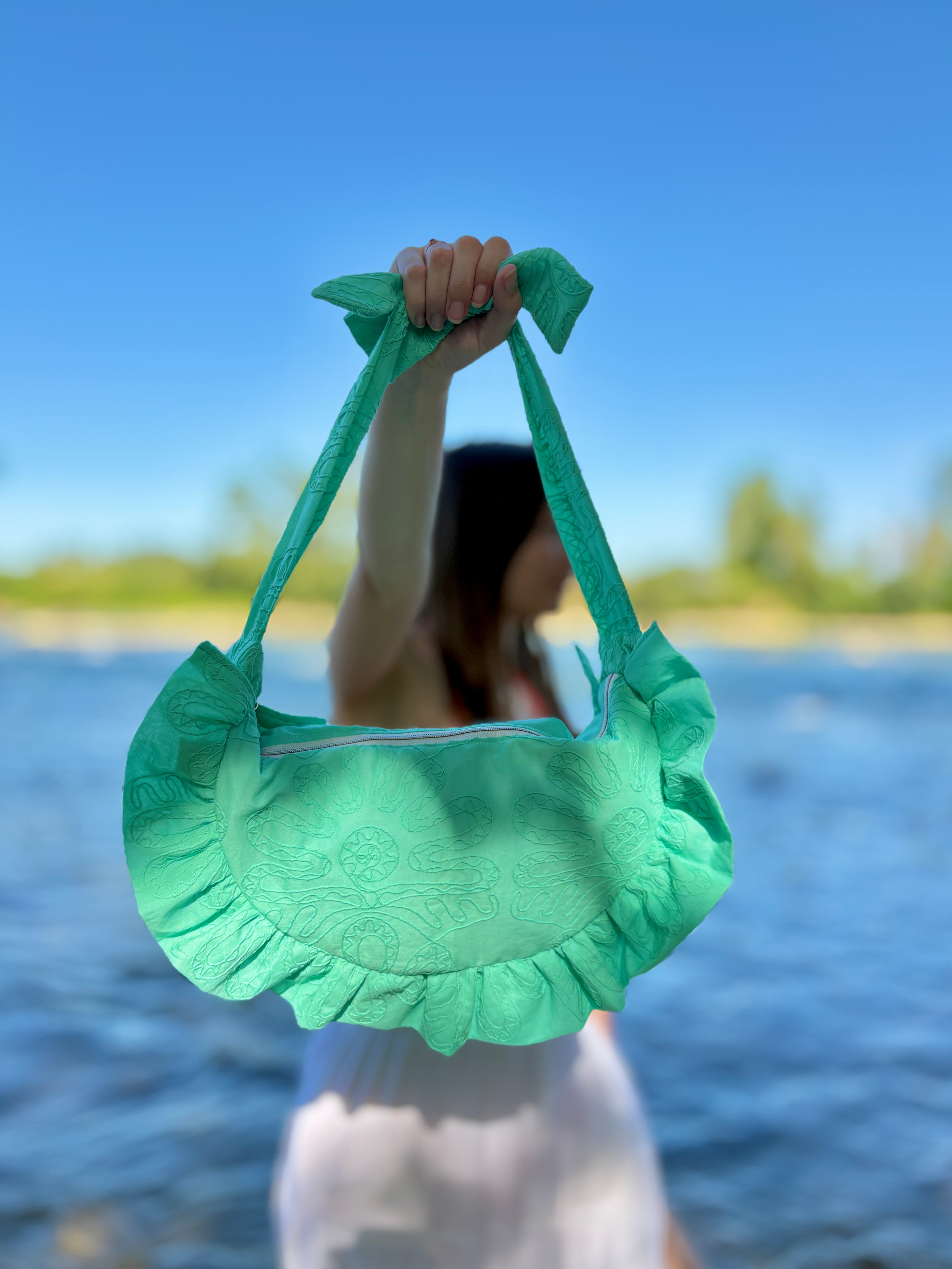 Mise en avant en mode portrait du sac cousu main de la marque SIOU en forme de croissant avec des volants au coloris lagon, un turquoise lumineux avec des broderies organiques.