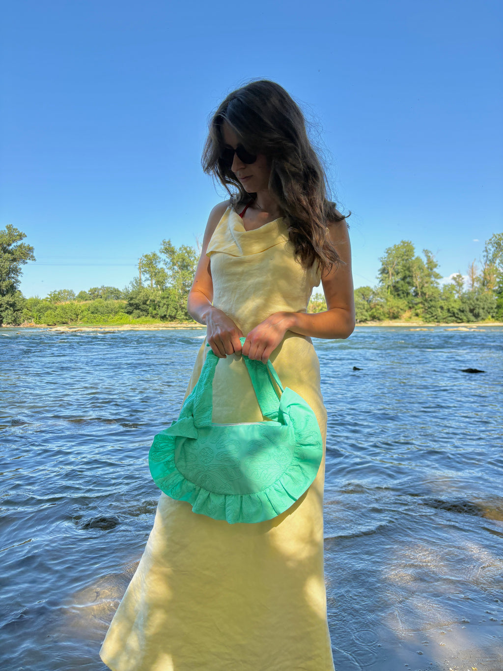 Femme habillée d'une robe longue jaune pastel au bord de l'eau sur la plage portant à ses mains le sac fait main Lagon de la marque SIOU, un coloris turquoise vif aux broderies fleuries.