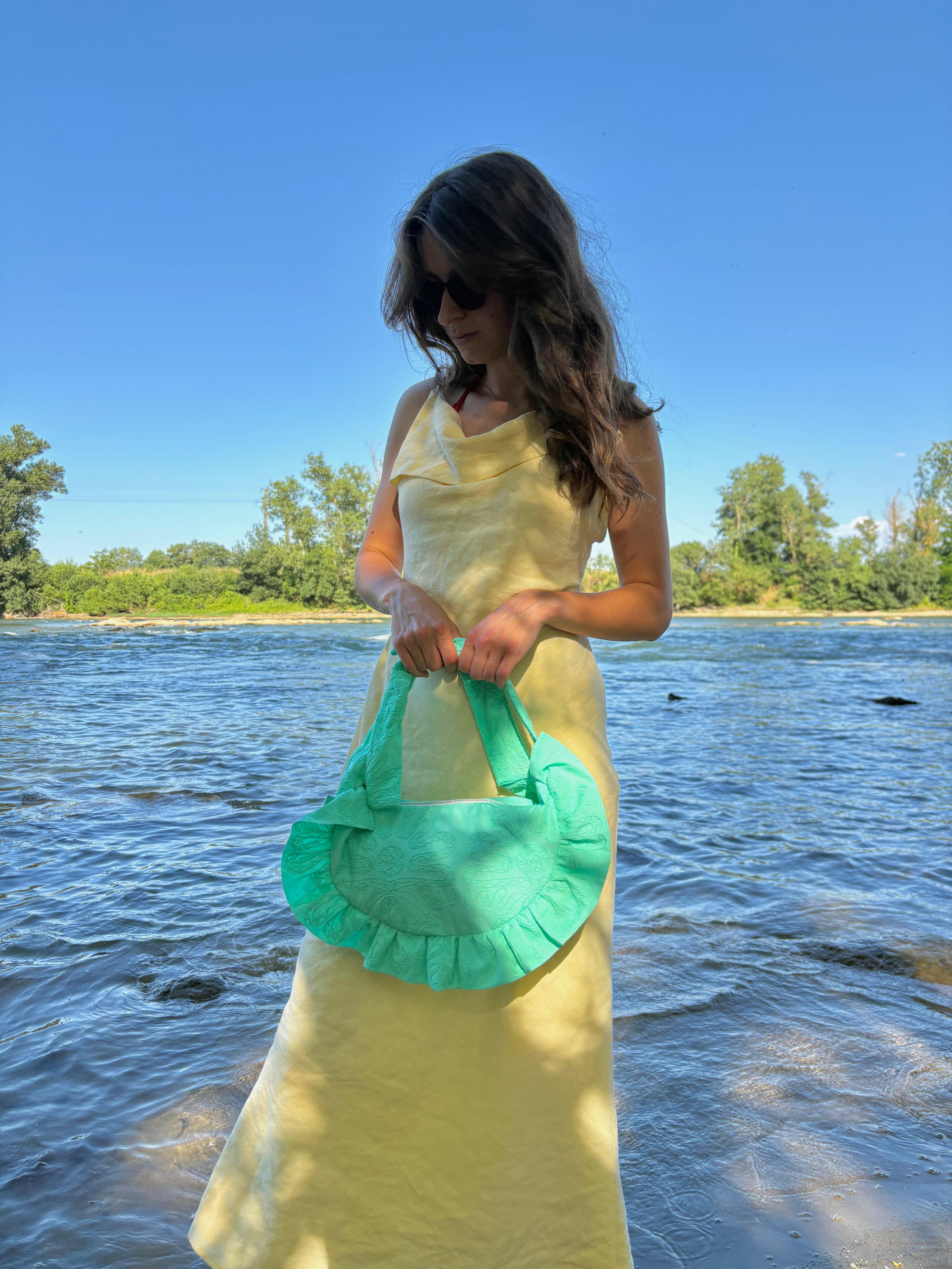 Femme habillée d'une robe longue jaune pastel au bord de l'eau sur la plage portant à ses mains le sac fait main Lagon de la marque SIOU, un coloris turquoise vif aux broderies fleuries.
