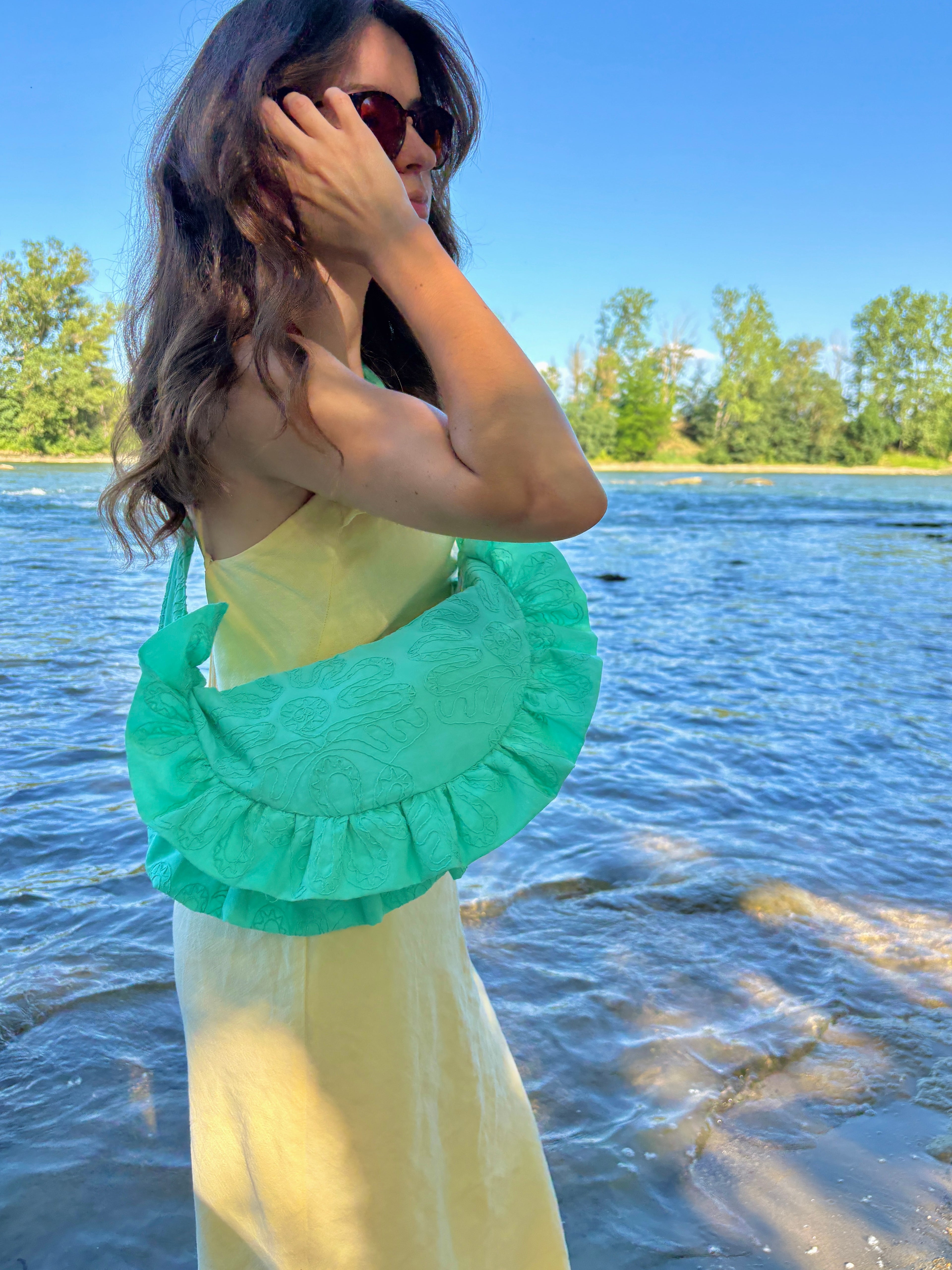 Jeune femme portant le sac cousu main SIOU Lagon en bandoulière. Le sac à la forme arrondie arborant des froufrous est de couleur bleu/vert turquoise flashy, avec des broderies fleuries. la femme se recoiffe légèrement au bord de l'eau.