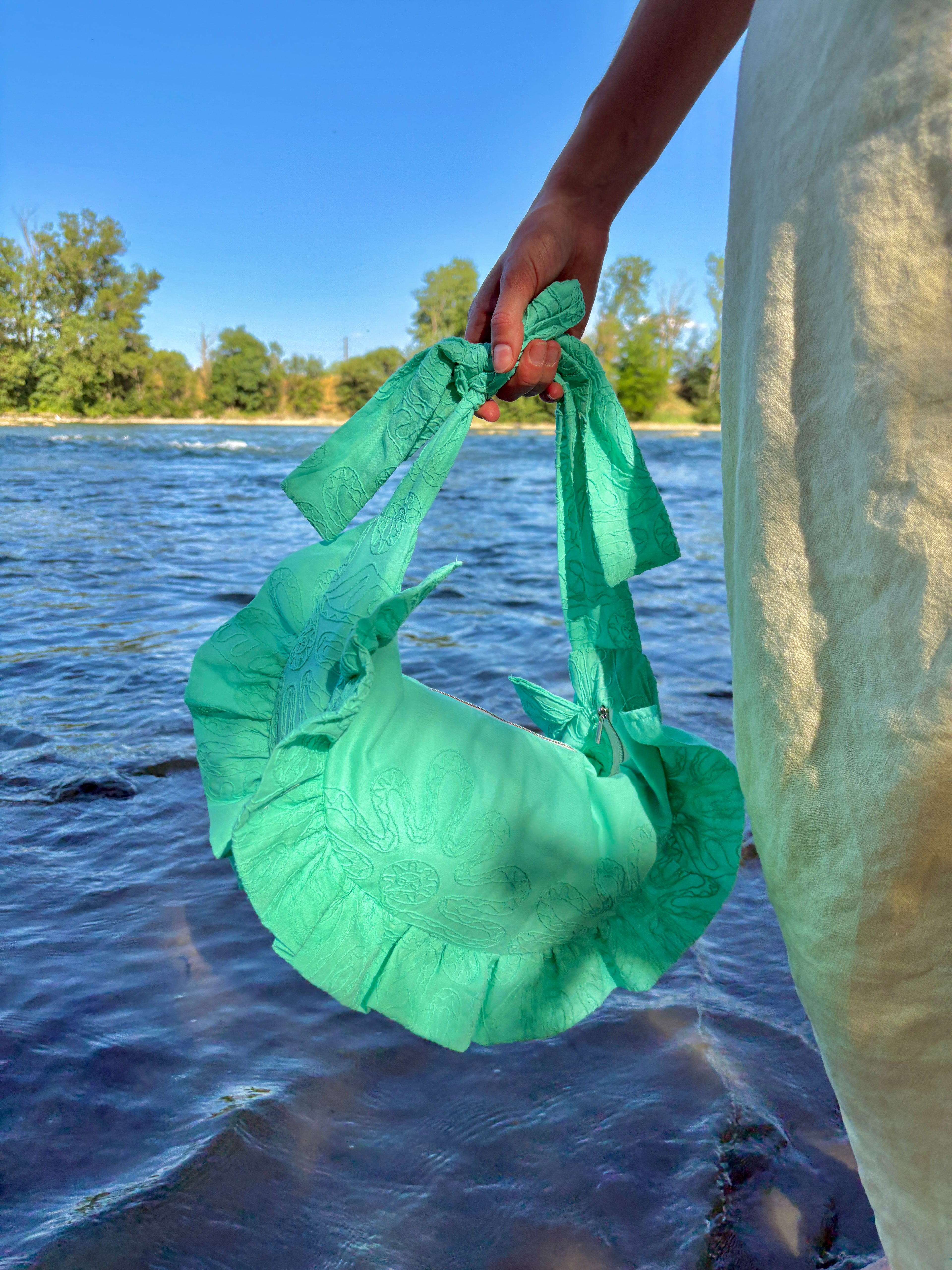 Sac à main en forme de croissant à volants coloris Lagon, un turquoise lumineux avec des broderies fleuries, de la marque SIOU, porté à la main en mouvement au bord de l'eau sur le sable d'une plage.