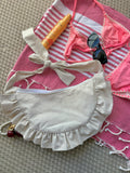 Sac à main Sable de la marque SIOU en tissu à volants en forme de demi-lune/croissant, cousu main, coloris lin beige, collection Été, sur une serviette de plage entouré d'un maillot de bain deux pièces rose, une crème solaire et des lunettes de soleil.