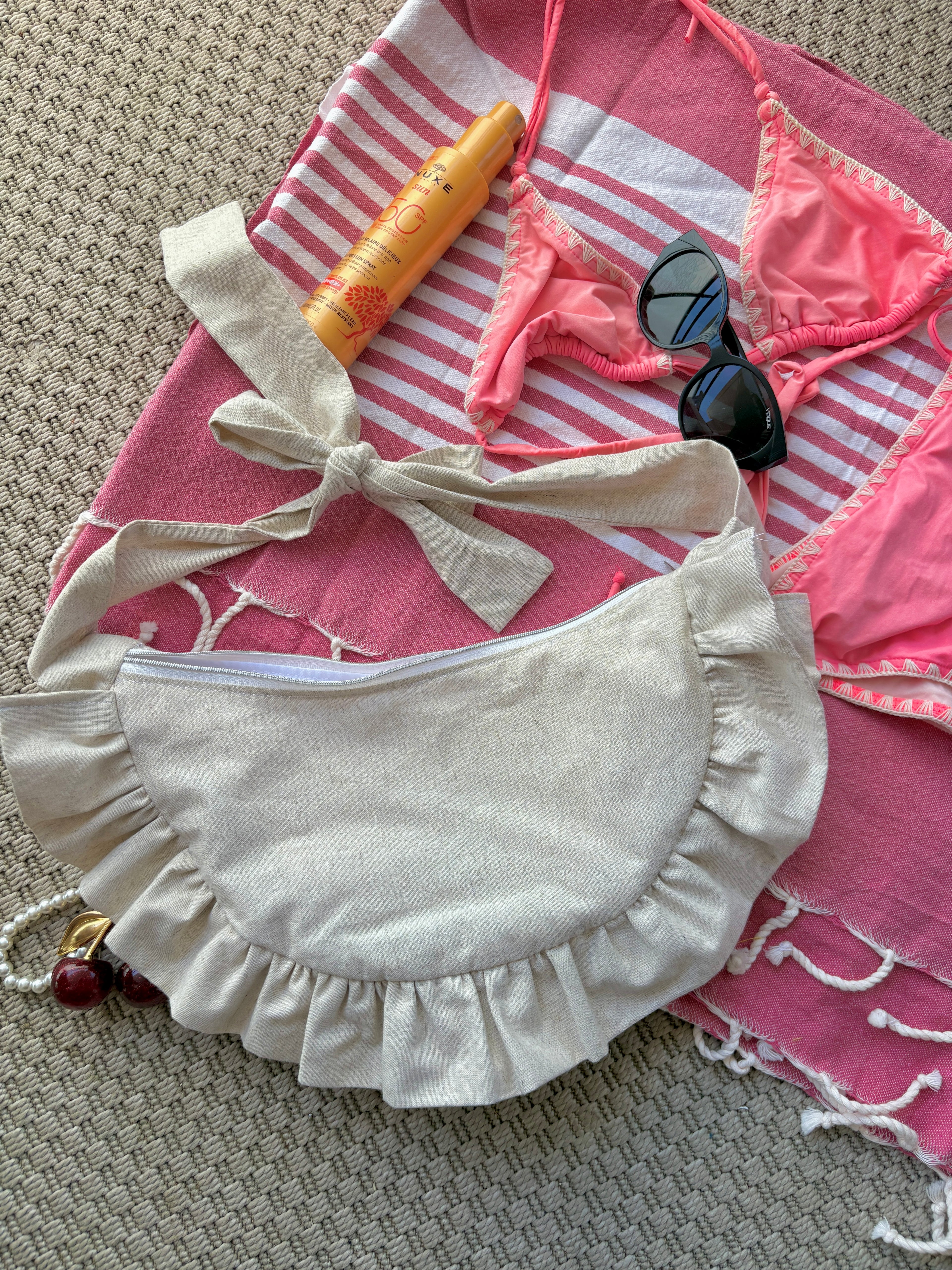 Sac à main Sable de la marque SIOU en tissu à volants en forme de demi-lune/croissant, cousu main, coloris lin beige, collection Été, sur une serviette de plage entouré d'un maillot de bain deux pièces rose, une crème solaire et des lunettes de soleil.