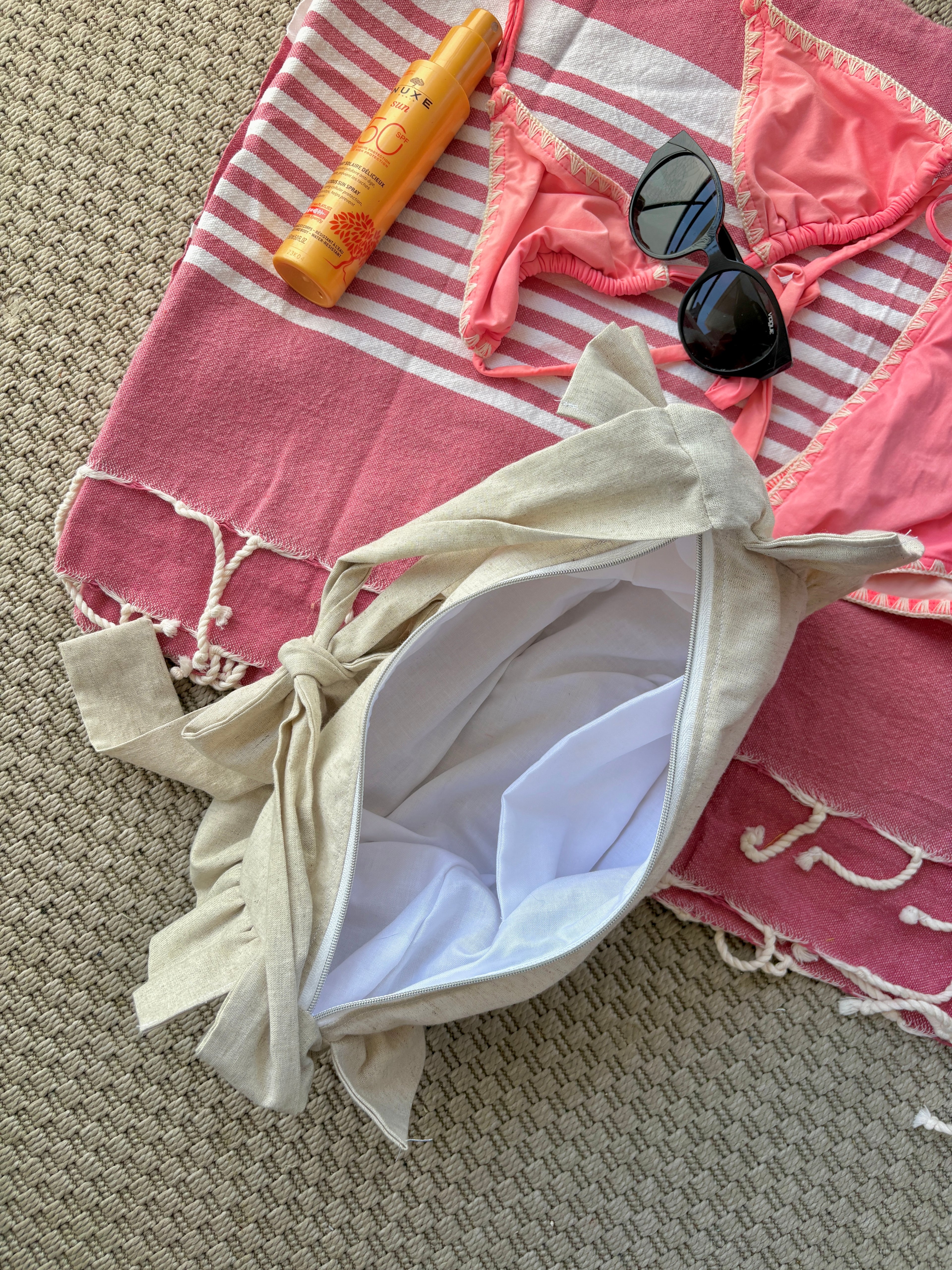 Vue du dessus de l'intérieur du sac à main Sable de la marque SIOU en tissu à volants en forme de demi-lune/croissant, cousu main, coloris lin beige, collection Été, sur une serviette de plage entouré d'un maillot de bain deux pièces rose, une crème solaire et des lunettes de soleil.