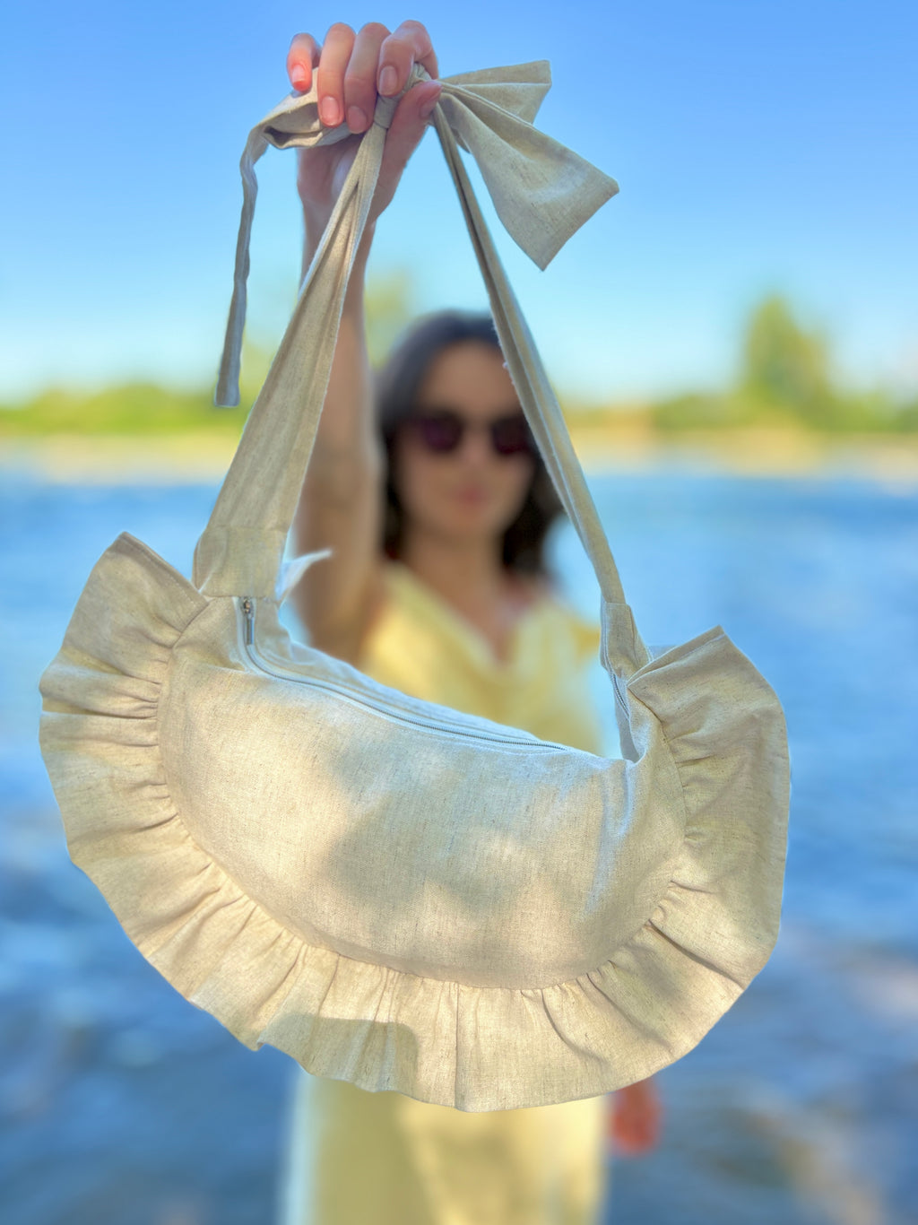 Mise en avant du sac Sable de la marque SIOU. Le sac, en forme de demi-lune à volants, de couleur beige en lin, tenu à bout de bras par un personne sur la plage en bord de mer, dans l'arrière plan qui est flou.