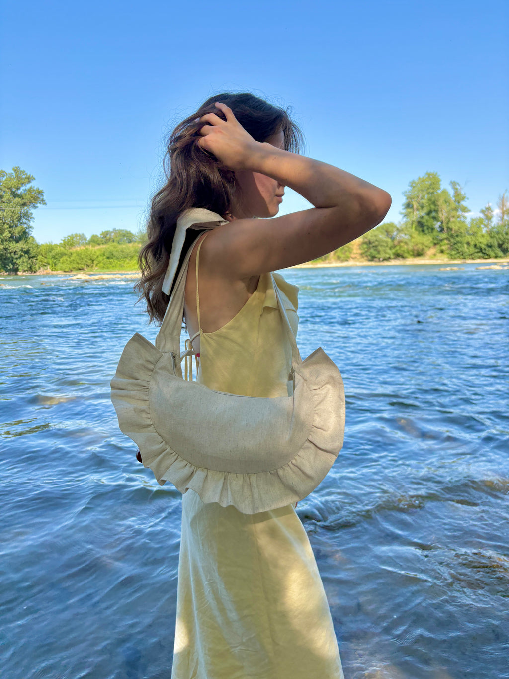 Jeune femme portant une robe jaune pastel se baladant sur la plage au bord de l'eau se recoiffant. elle porte à l'épaule le sac demi-lune cousu main à volants Sable de la marque SIOU, un coloris uni beige lin.