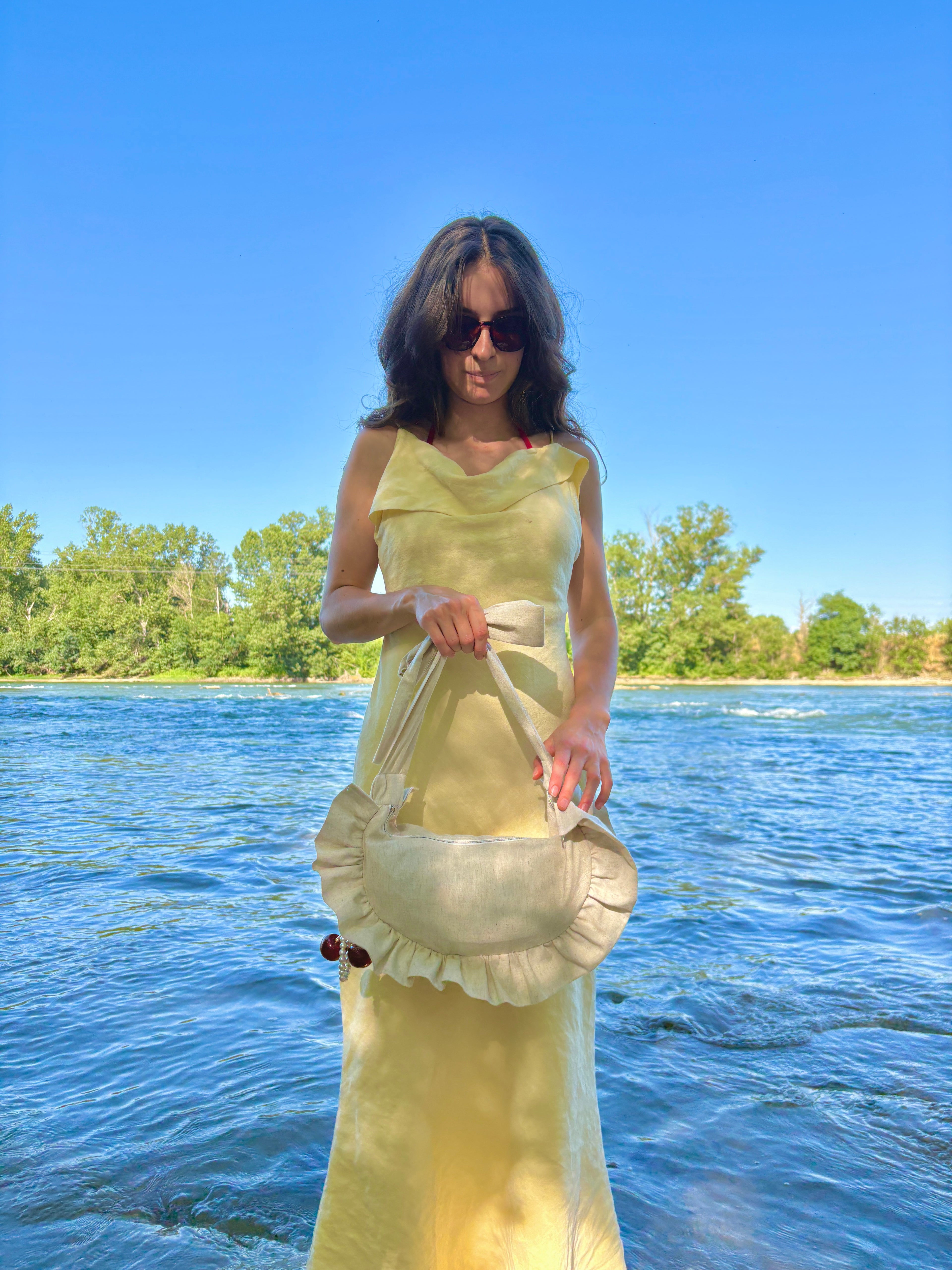 Au bord de l'eau, sur la plage, une femme se tient debout, de face, elle est vêtue d'une longue robe jaune et pote à la main le sac à main en forme de croissant à volants Sable, de la marque SIOU, c'est un sac cousu main, il est de couleur beige uni.
