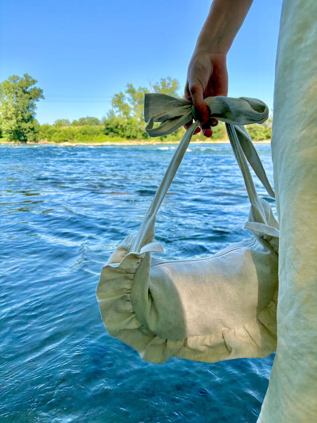 Sac à main demi-lune-/croissant Sable de la marque SIOU porté à la main au bord de l'eau. C'est un sac fait main dont les anses peuvent être nouées à la hauteur souhaitée pour le porter soit à l'épaule, soit à la main, soit en bandoulière.