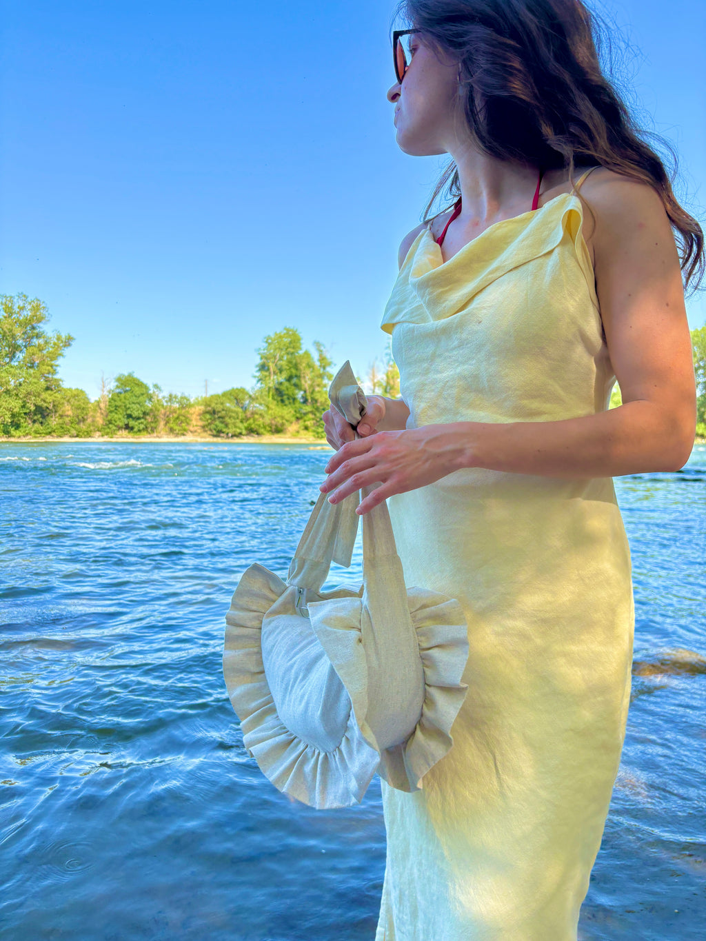 Jeune femme sur la plage au bord de la mer, portant une robe jaune pastel porte à ses mains le sac Sable de la marque SIOU. le sac est de couleur beige lin, en forme de demi-lune à froufrous et est entièrement fait main.
