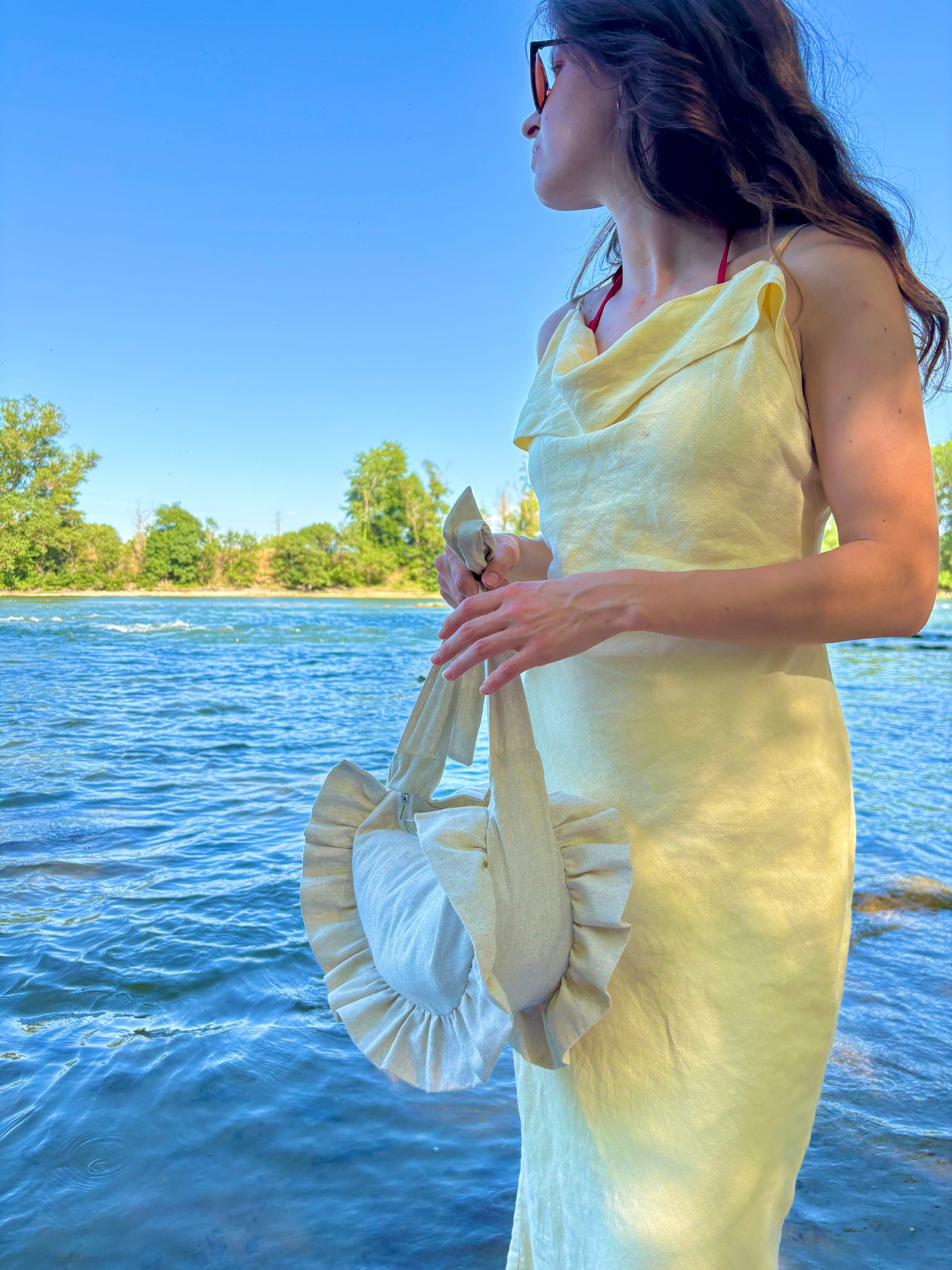 Jeune femme sur la plage au bord de la mer, portant une robe jaune pastel porte à ses mains le sac Sable de la marque SIOU. le sac est de couleur beige lin, en forme de demi-lune à froufrous et est entièrement fait main.