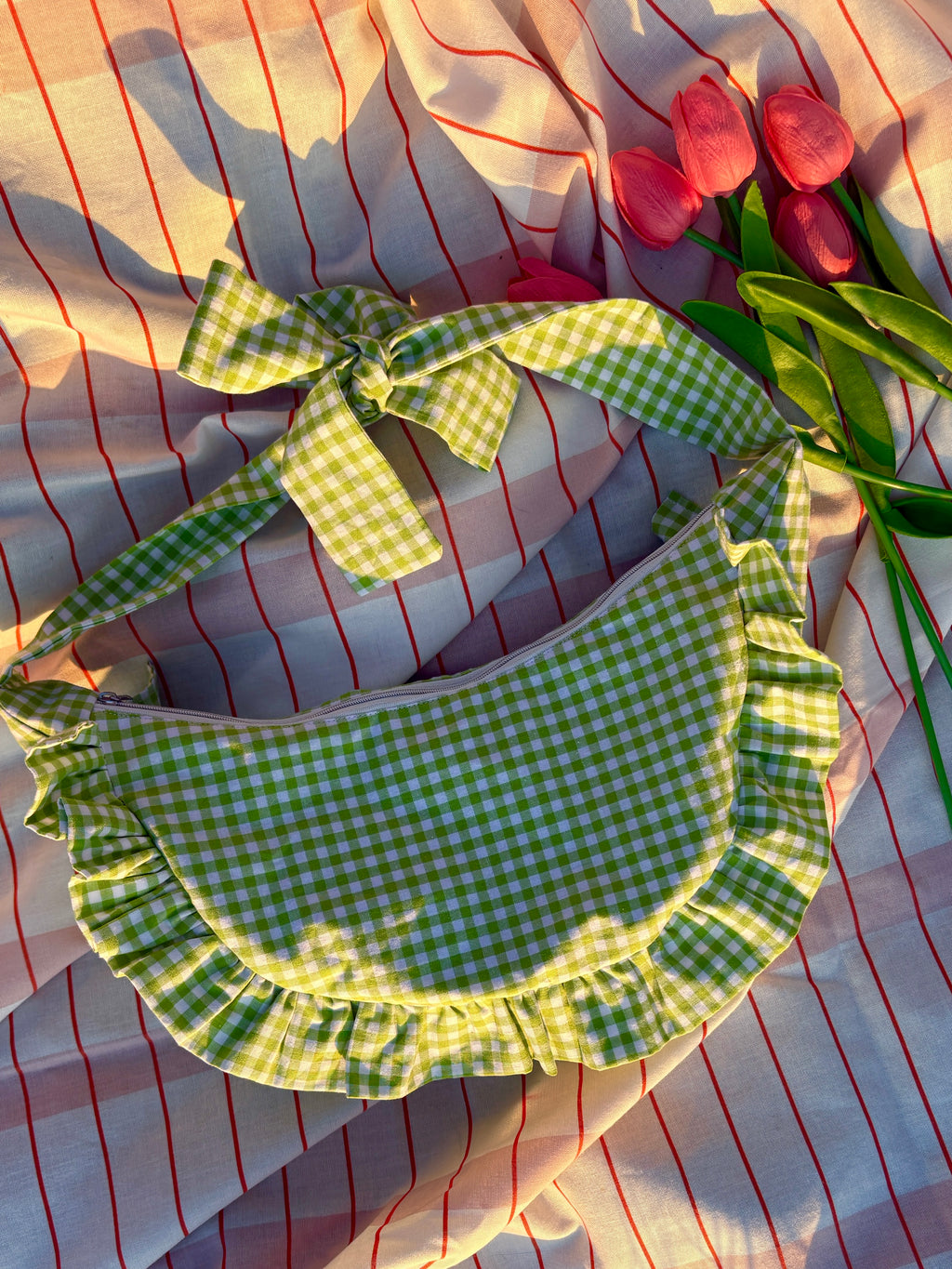 Sac à main en tissu à volants en forme de demi-lune/croissant, cousu main, coloris en vichy vert et blanc Basilic, collection Printemps, sur une nappe de pique-nique, sur de l'herbe avec des tulipes autour du sac de la marque SIOU
