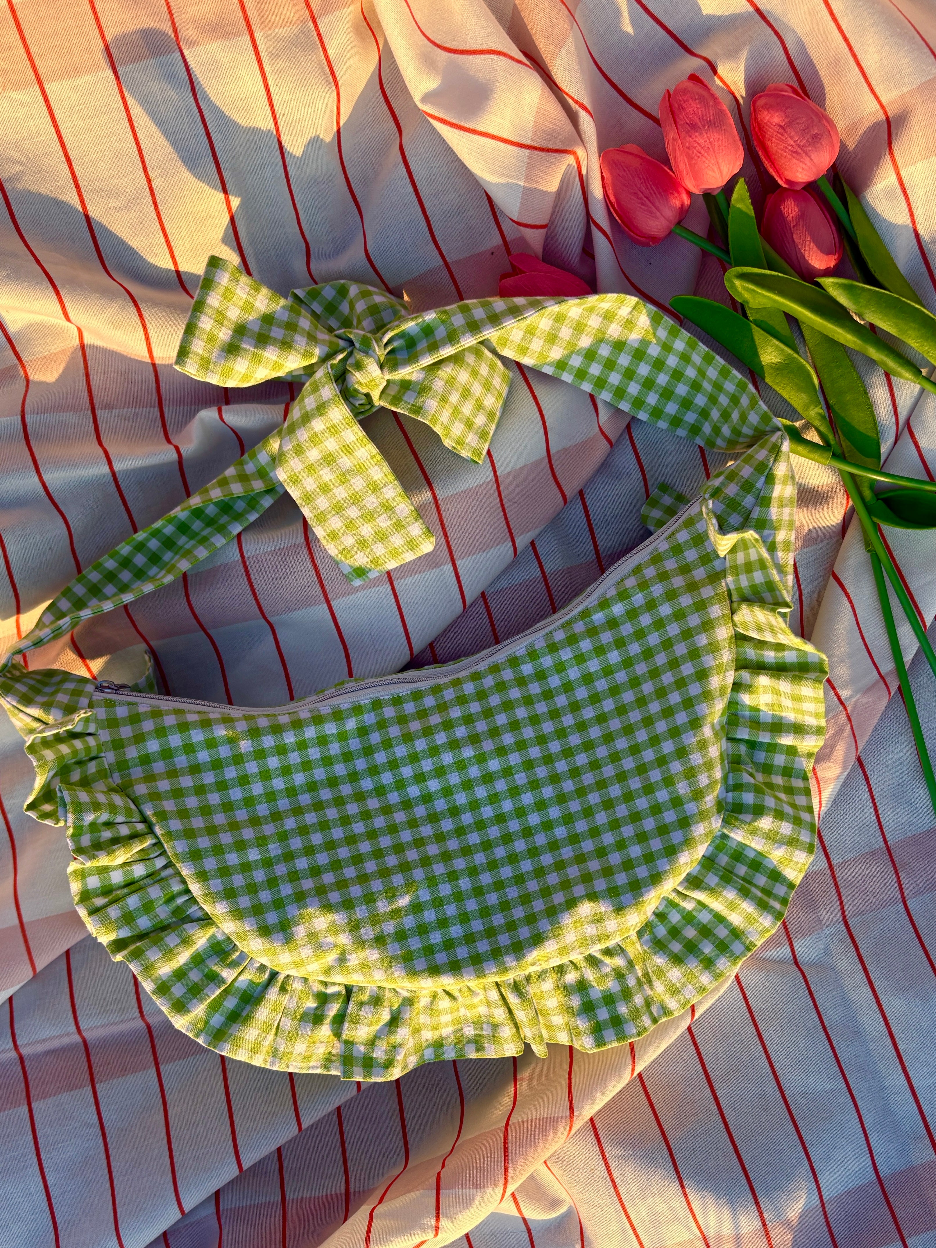 Sac à main en tissu à volants en forme de demi-lune/croissant, cousu main, coloris en vichy vert et blanc Basilic, collection Printemps, sur une nappe de pique-nique, sur de l'herbe avec des tulipes autour du sac de la marque SIOU