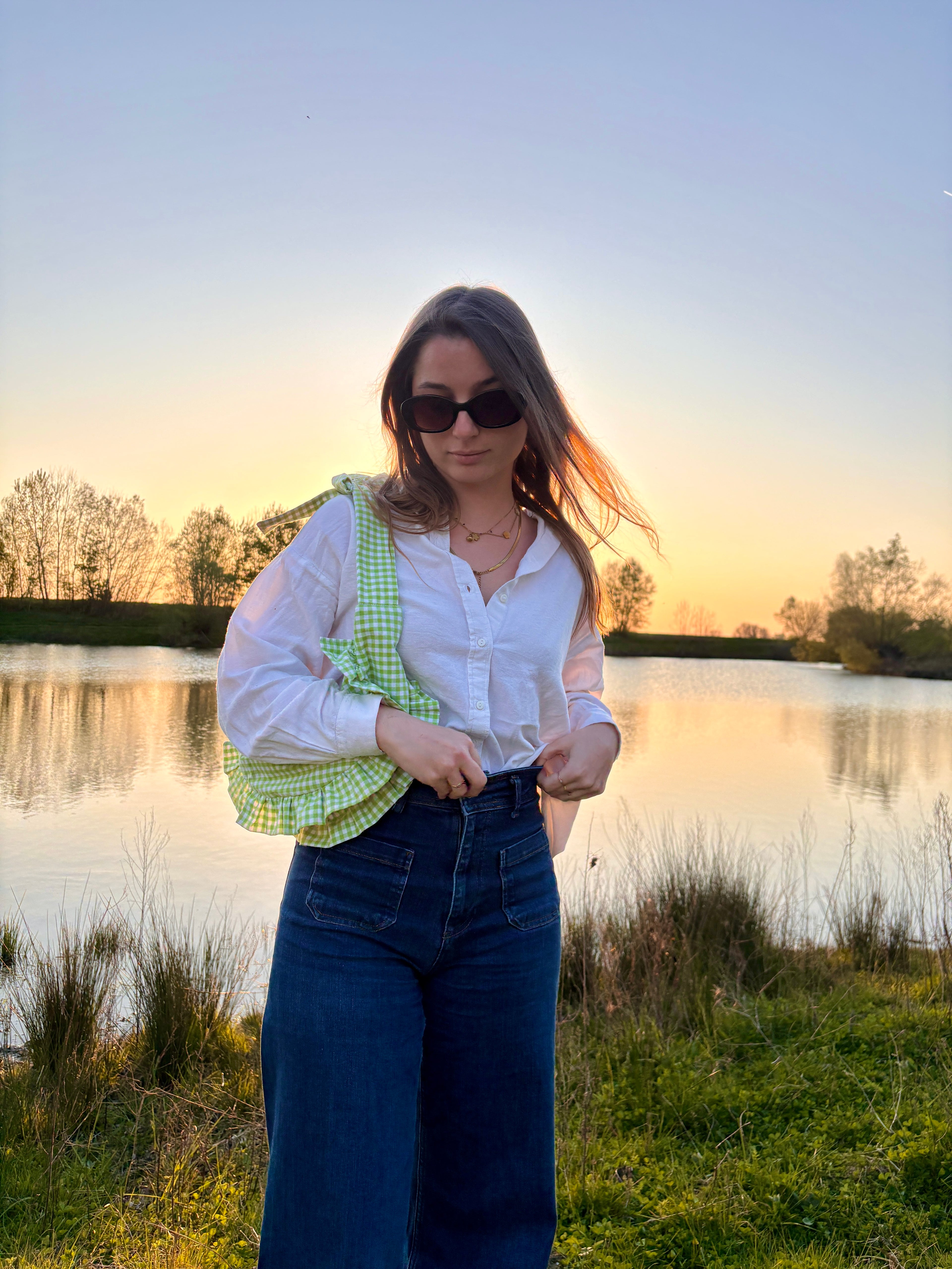 Sac à main SIOU en tissu à volants en forme de demi-lune/croissant, fait main, coloris en vichy vert et blanc Basilic, collection Printemps. Il est porté à l'épaule par une femme en chemise blanche et jean au bord d'un grand lac au coucher du soleil