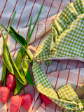 Plan rapproché sur les détails et coutures du sac à main en tissu à volants en forme de demi-lune/croissant, cousu main, coloris en vichy vert et blanc Basilic, collection Printemps, sur une nappe de pique-nique, avec des tulipes autour du sac de la marque SIOU