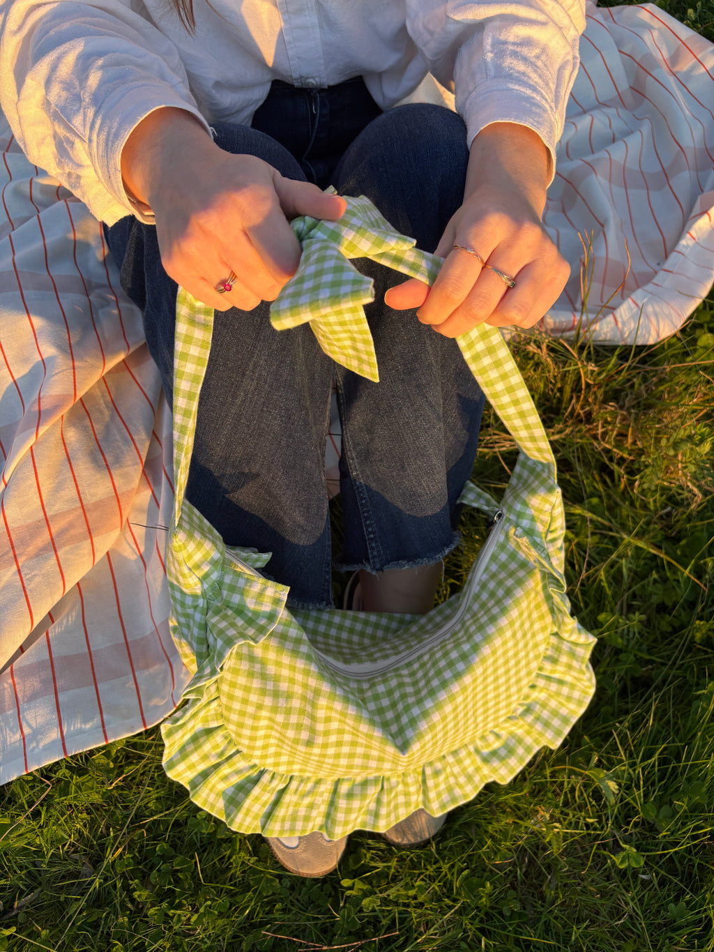 Sac à main en tissu à volants en forme de demi-lune/croissant, couture fait main, coloris en vichy vert et blanc Basilic, collection Printemps, porté à la main par une fille assise sur une nappe de pique-nique, sur de l'herbe.