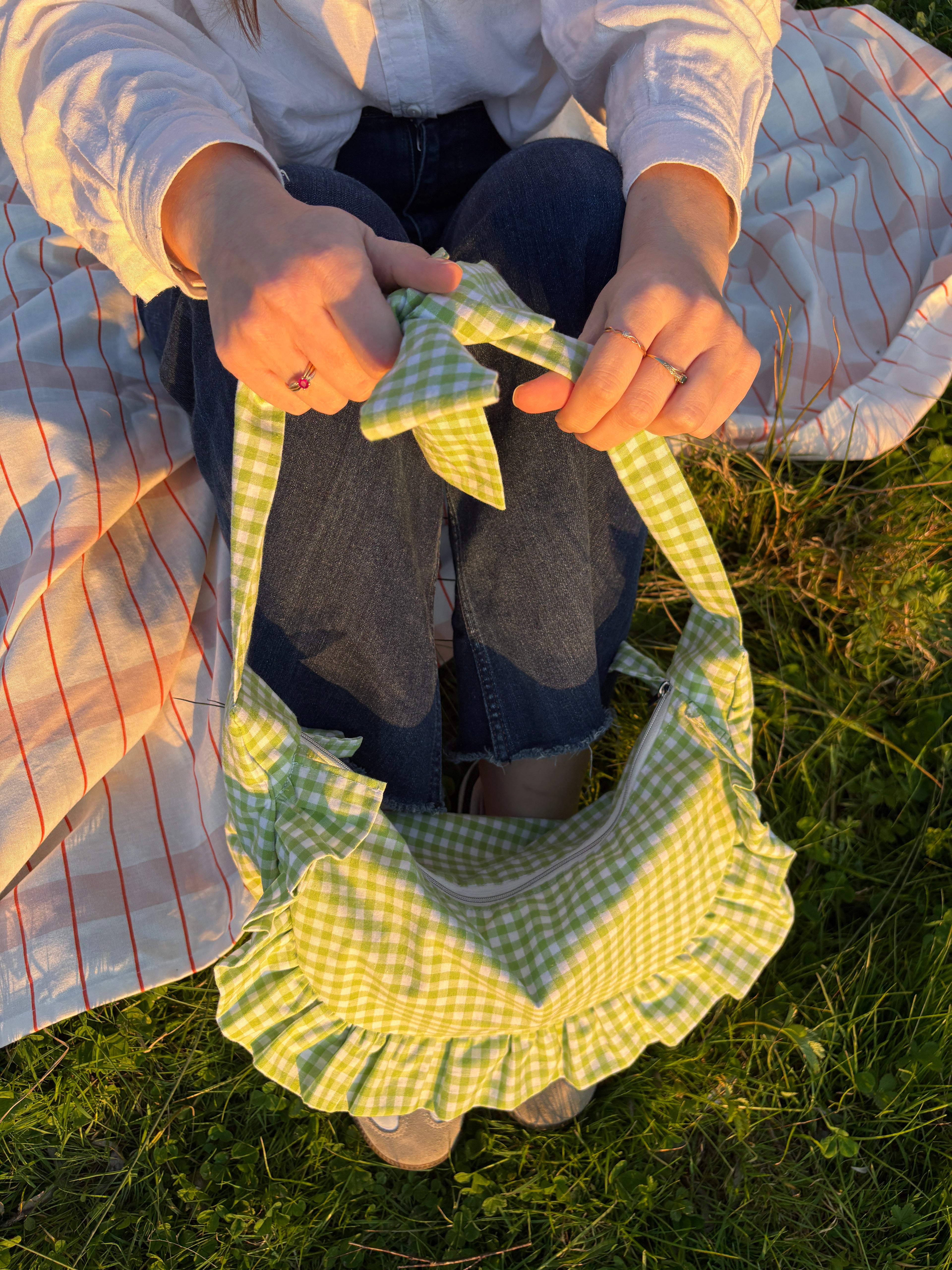 Sac à main en tissu à volants en forme de demi-lune/croissant, couture fait main, coloris en vichy vert et blanc Basilic, collection Printemps, porté à la main par une fille assise sur une nappe de pique-nique, sur de l'herbe.