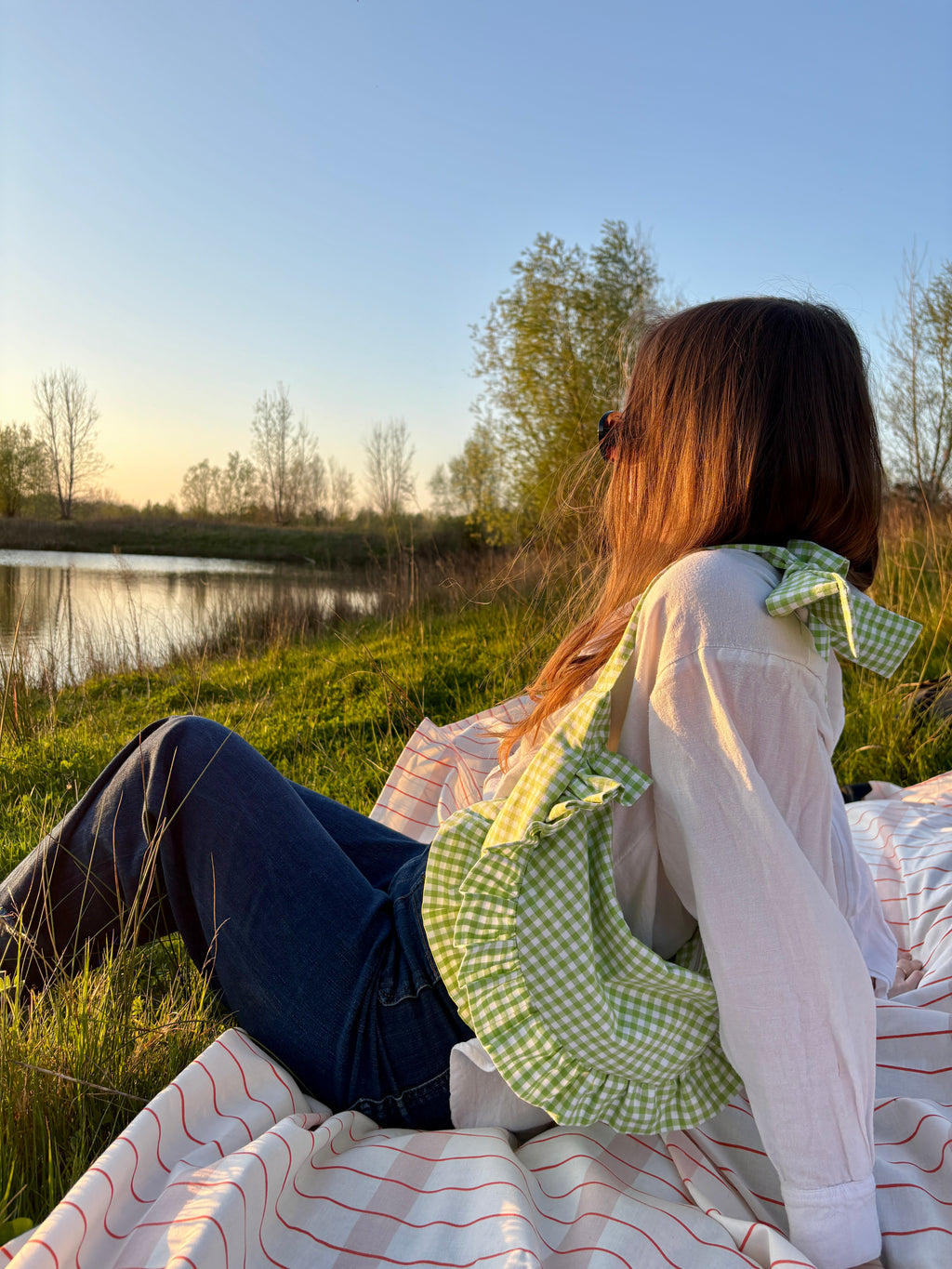 Sac à main de la marque SIOU en tissu à volants en forme de demi-lune/croissant, artisanal, coloris en vichy vert et blanc Basilic, collection Printemps, porté épaule par une jeune femme assise de profil regardant un grand lac au coucher du soleil sur une nappe de pique-nique, sur de l'herbe.
