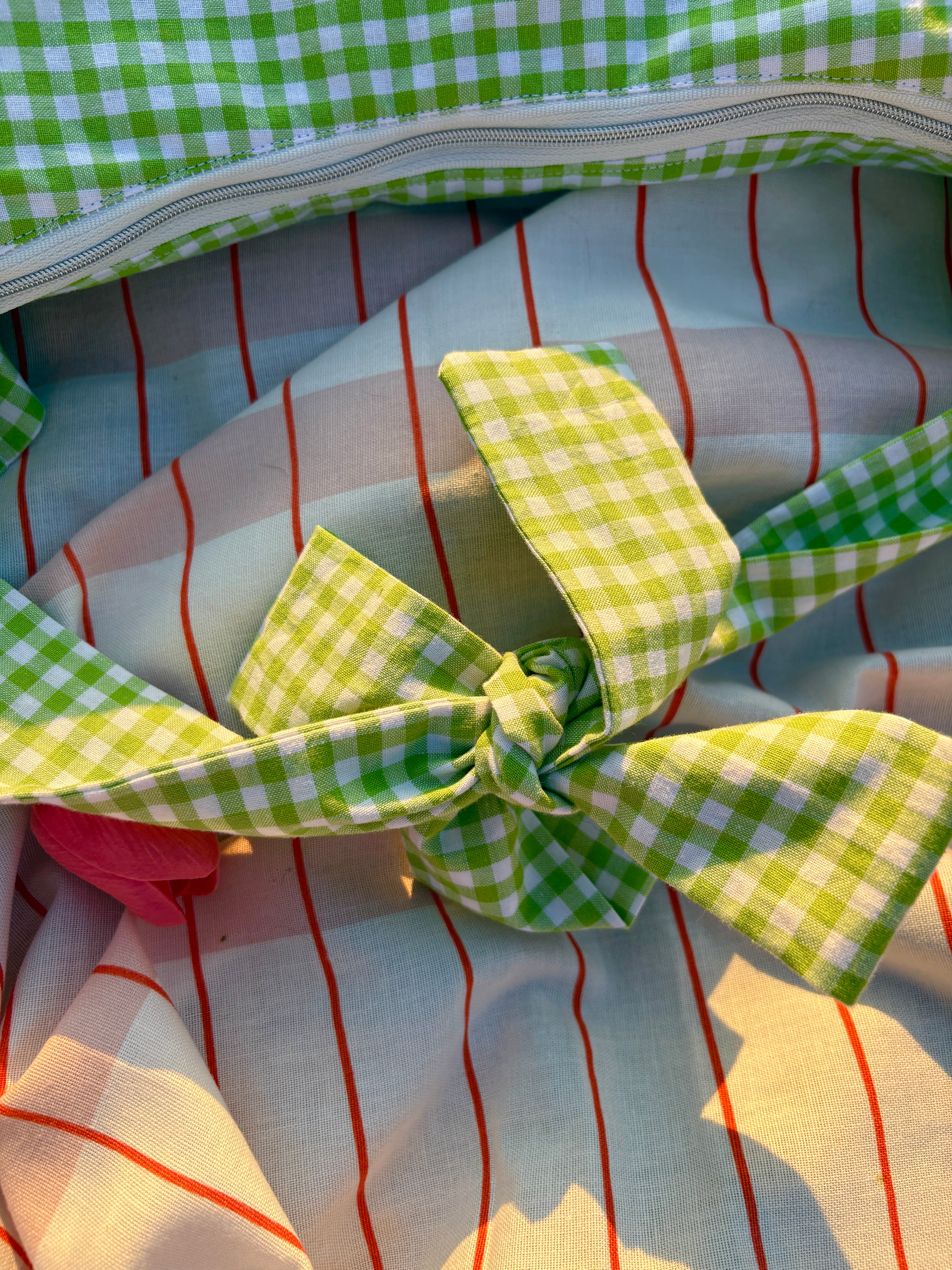 Zoom sur les détails de couture et du nœud du sac à main en tissu à volants en forme de croissant, cousu main, coloris en vichy vert et blanc Basilic, collection Printemps, sur une nappe de pique-nique, avec des tulipes autour du sac de la marque SIOU