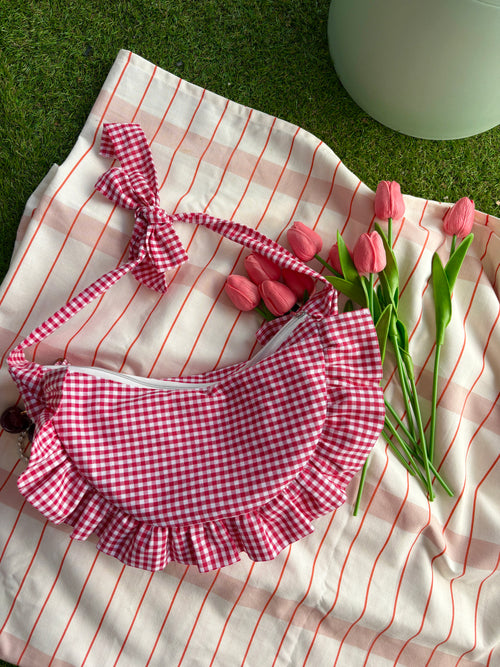Sac à main en tissu à volants en forme de demi-lune/croissant, cousu main, coloris en vichy rouge et blanc Fraise, collection Printemps, sur une nappe de pique-nique, sur de l'herbe avec des tulipes de la marque SIOU